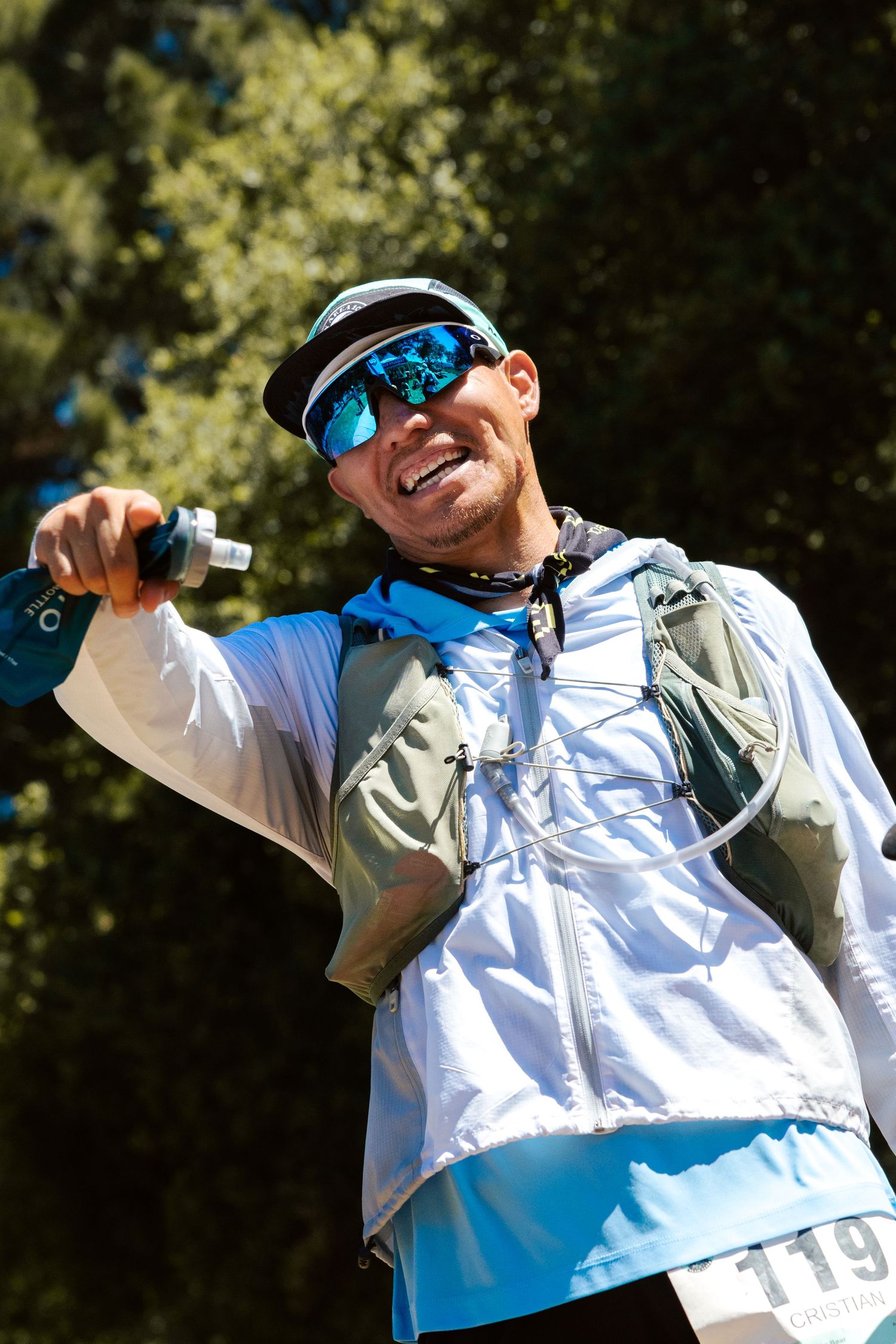 Smiling male trail runner wearing blue mirrored sunglasses, white jacket, and hydration vest, holding a water bottle outdoors.
