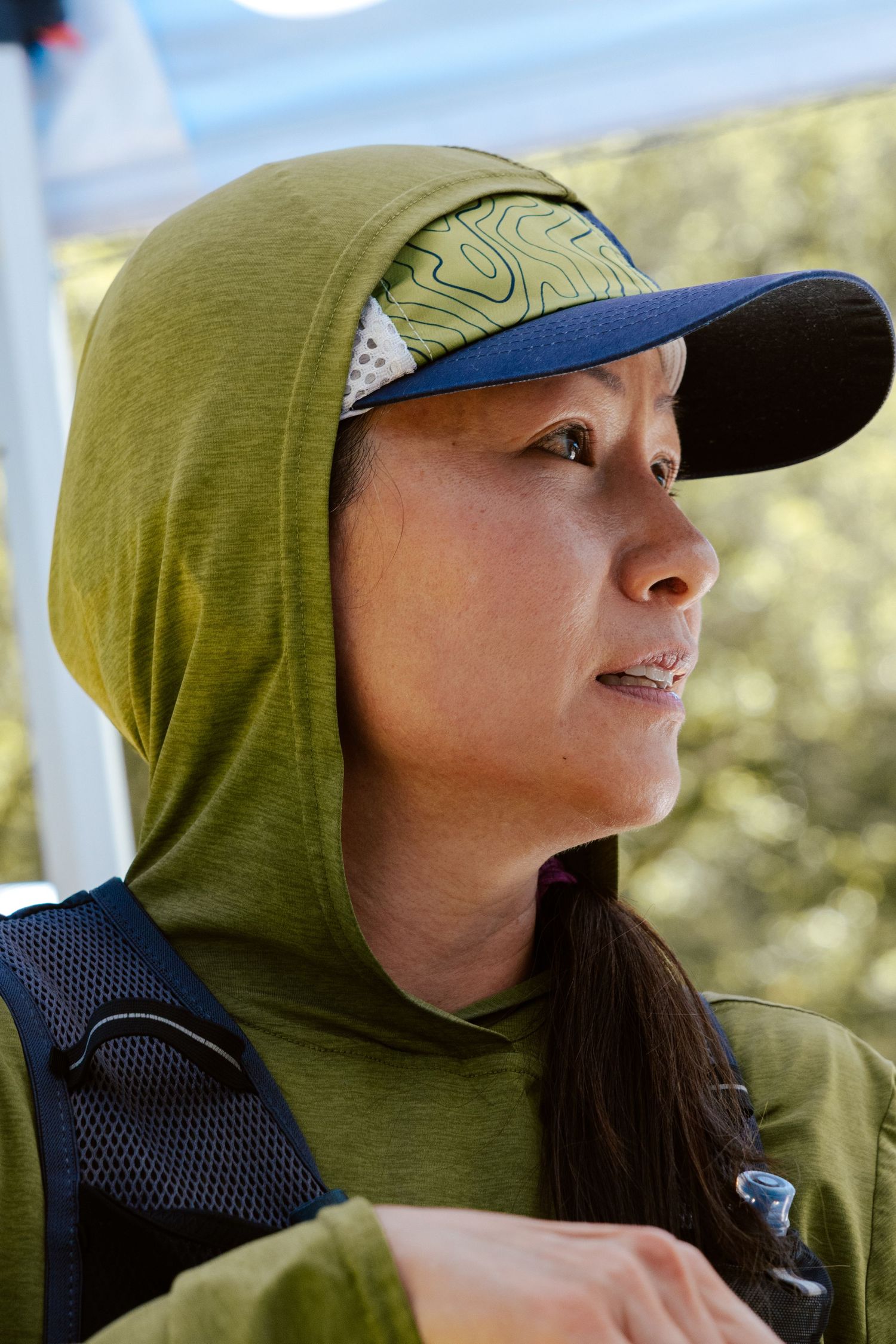Woman in green hoodie and blue cap with backpack gazing outdoors in natural light.
