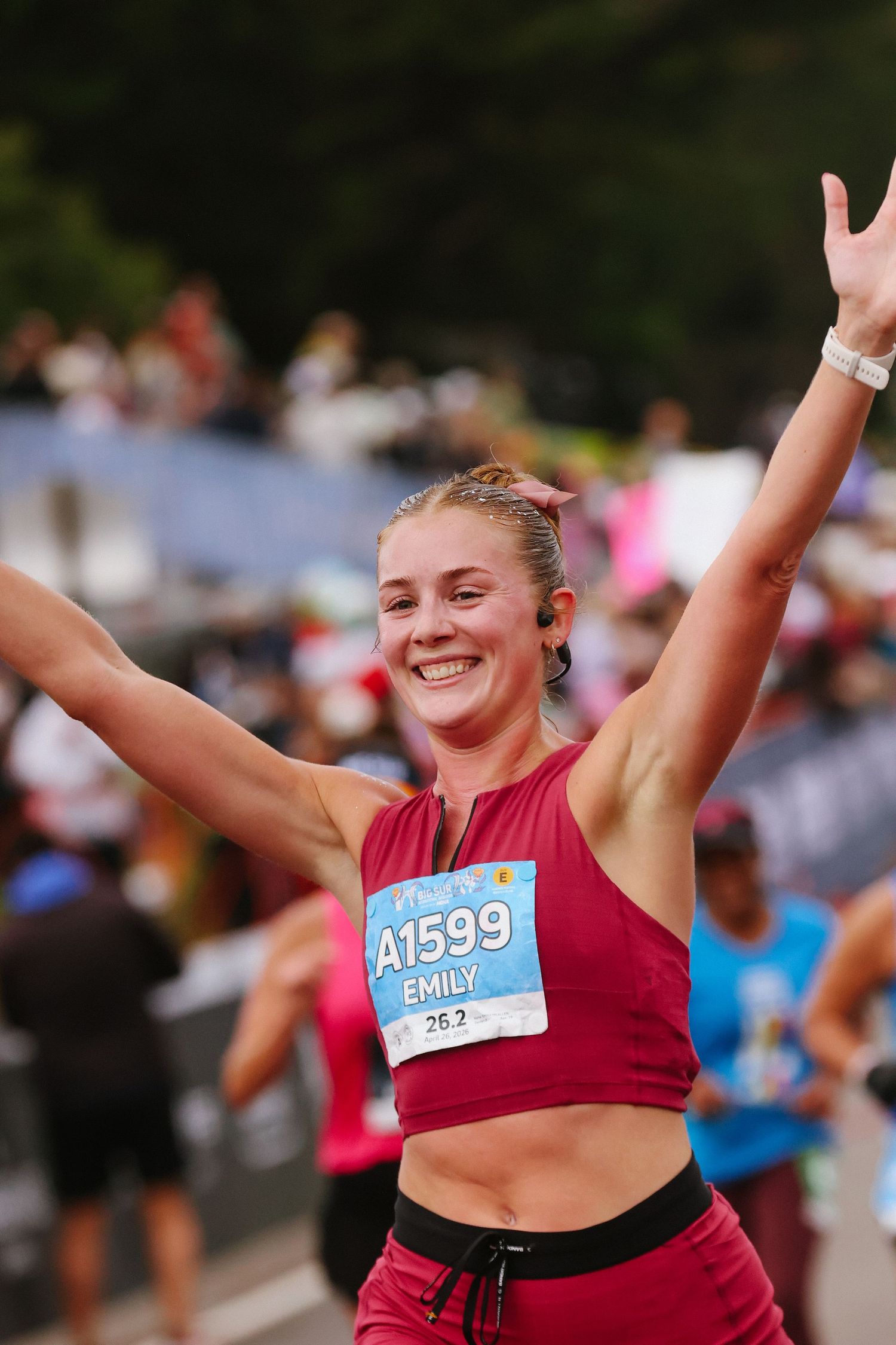 Smiling woman named Emily celebrates finishing a marathon with arms raised, wearing bib number A1599.