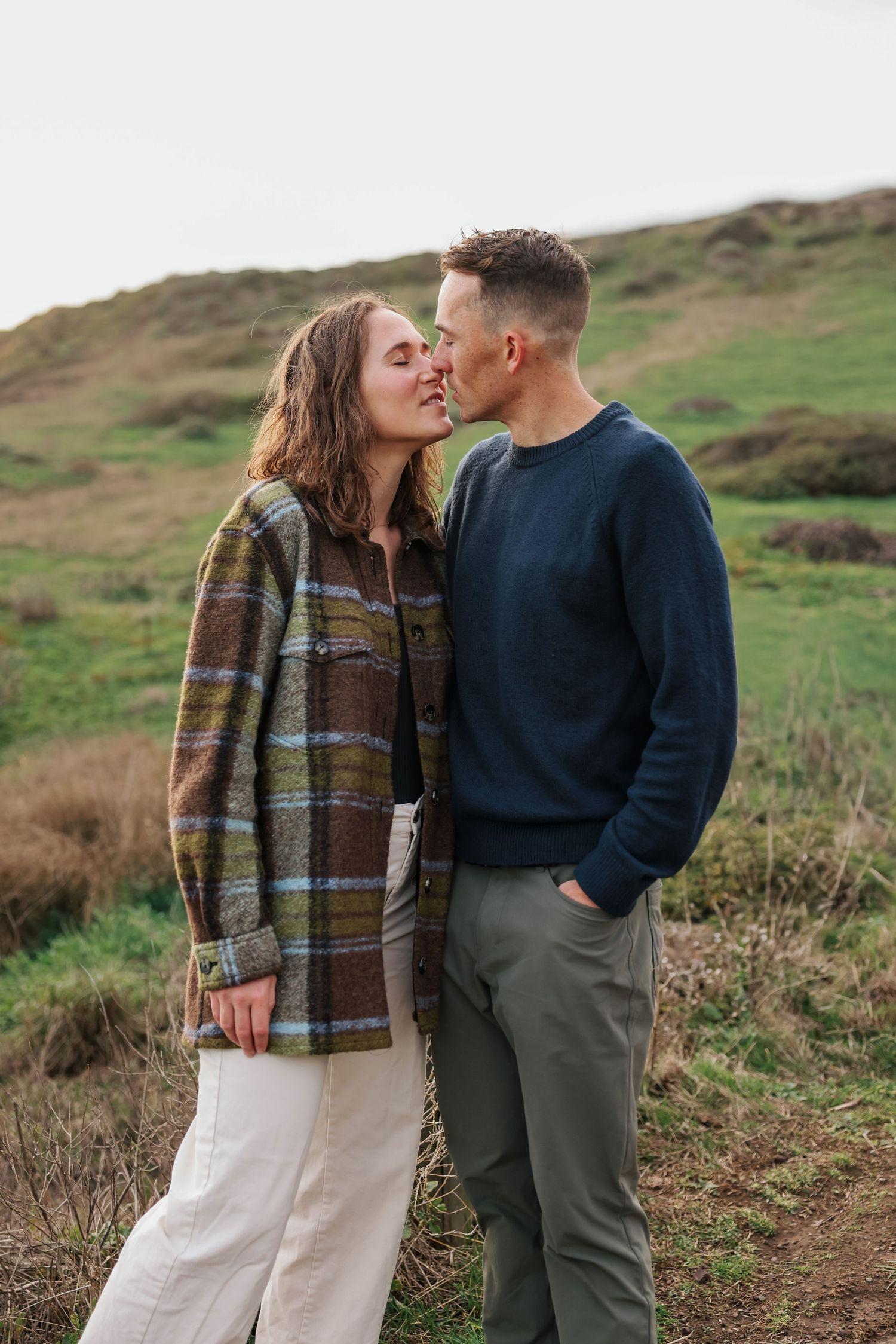 A couple sharing a kiss outdoors on a green hillside, woman in plaid coat and white pants, man in navy sweater.