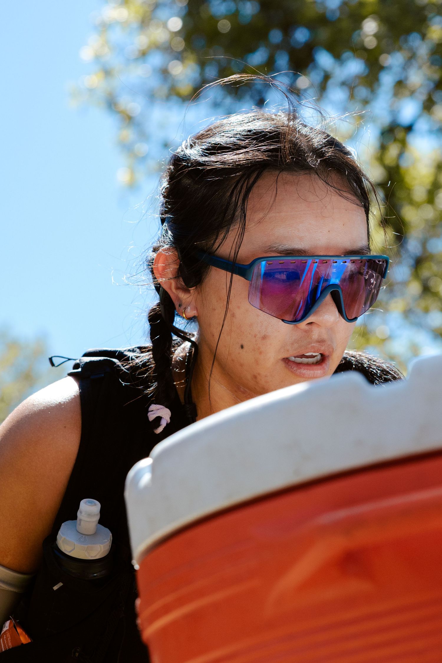 Young woman with blue mirrored sunglasses and braided hair outdoors on a sunny day near an orange barrel.