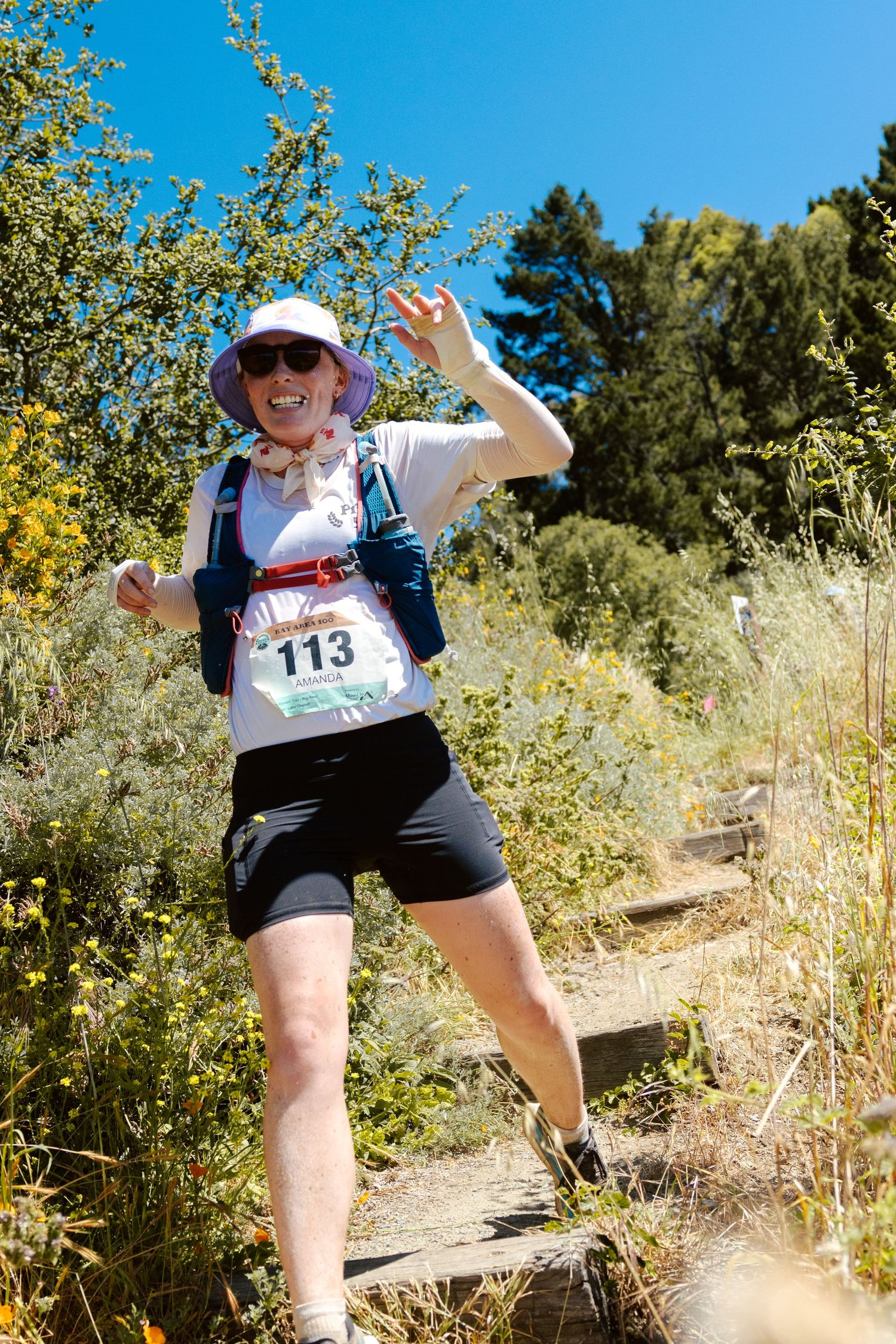 Smiling female trail runner wearing bib number 113 and a hat waves cheerfully on a sunny mountain trail.