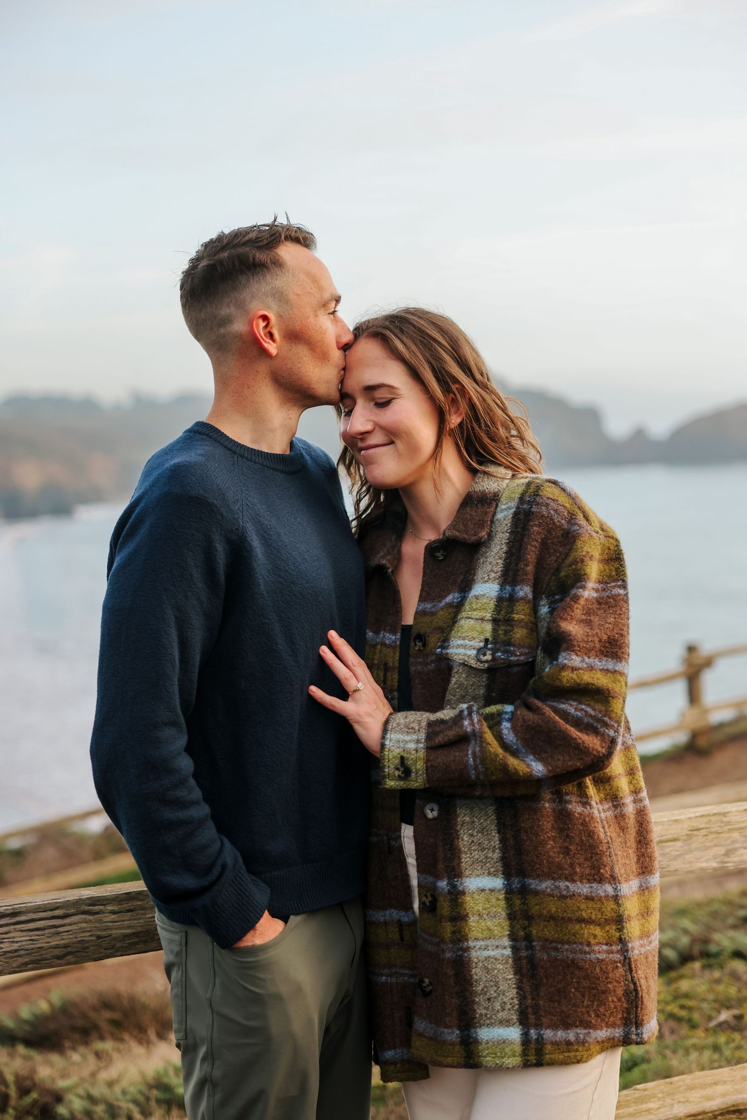 Man kissing woman on forehead near coastal fence, woman smiling and showing engagement ring on hand.