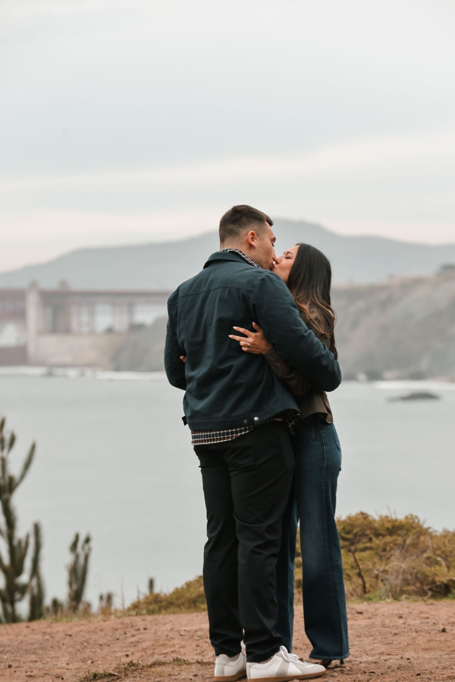 Newly engaged couple kissing on coastal overlook with Golden Gate Bridge and bay in background.