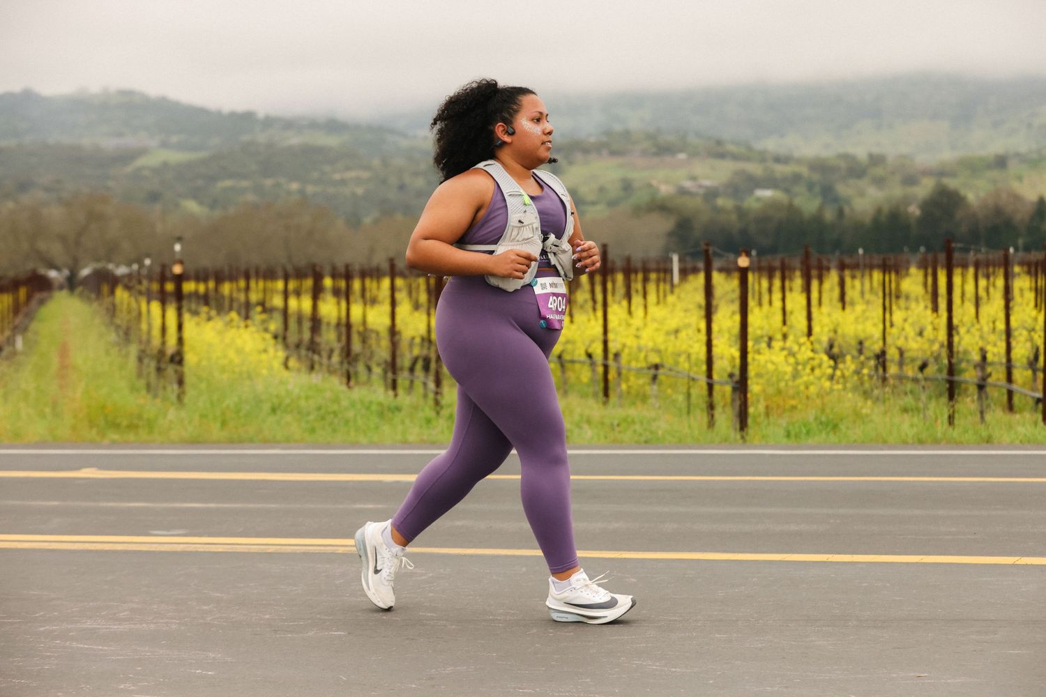 Female runner in purple leggings and vest jogs along a road past yellow wildflowers and vineyard hills.