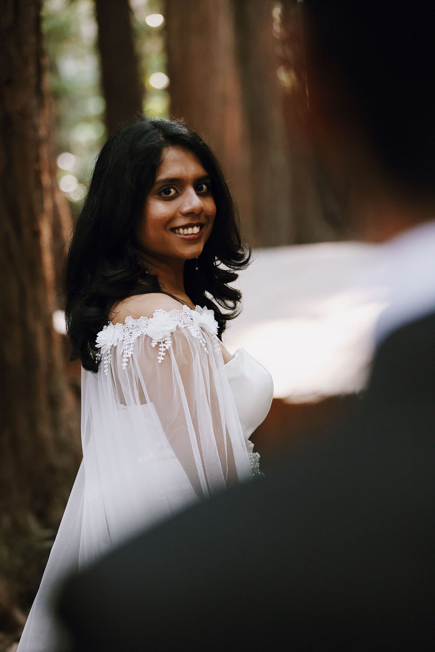 Indian bride in white off-shoulder lace wedding dress smiling over shoulder in forest setting.