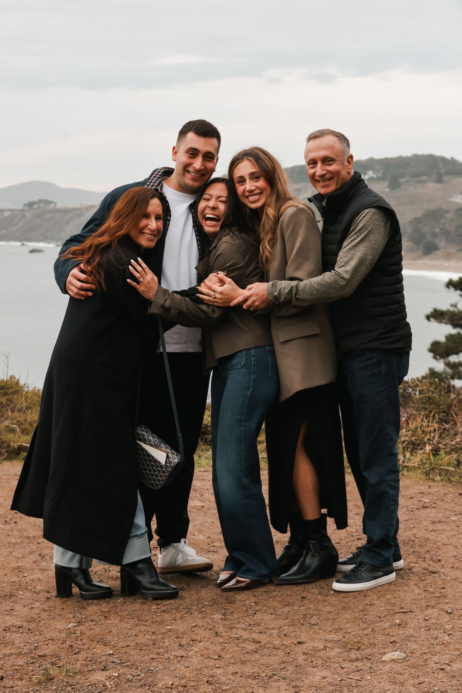 Five friends embrace and laugh together on a coastal cliff overlooking the ocean on a cloudy day.