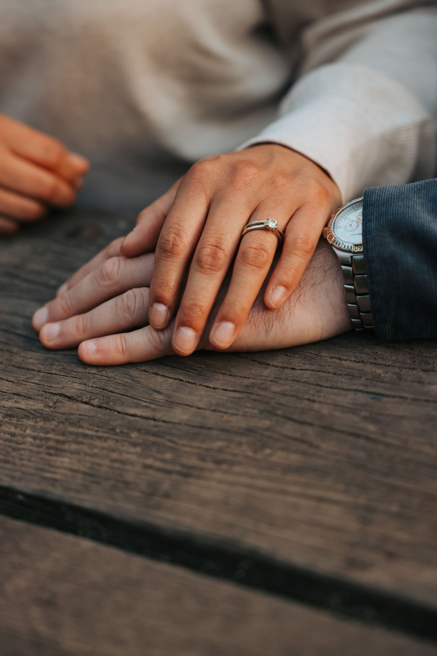 Close-up of engaged couples hands on a wooden table, showcasing a diamond engagement ring and watch.