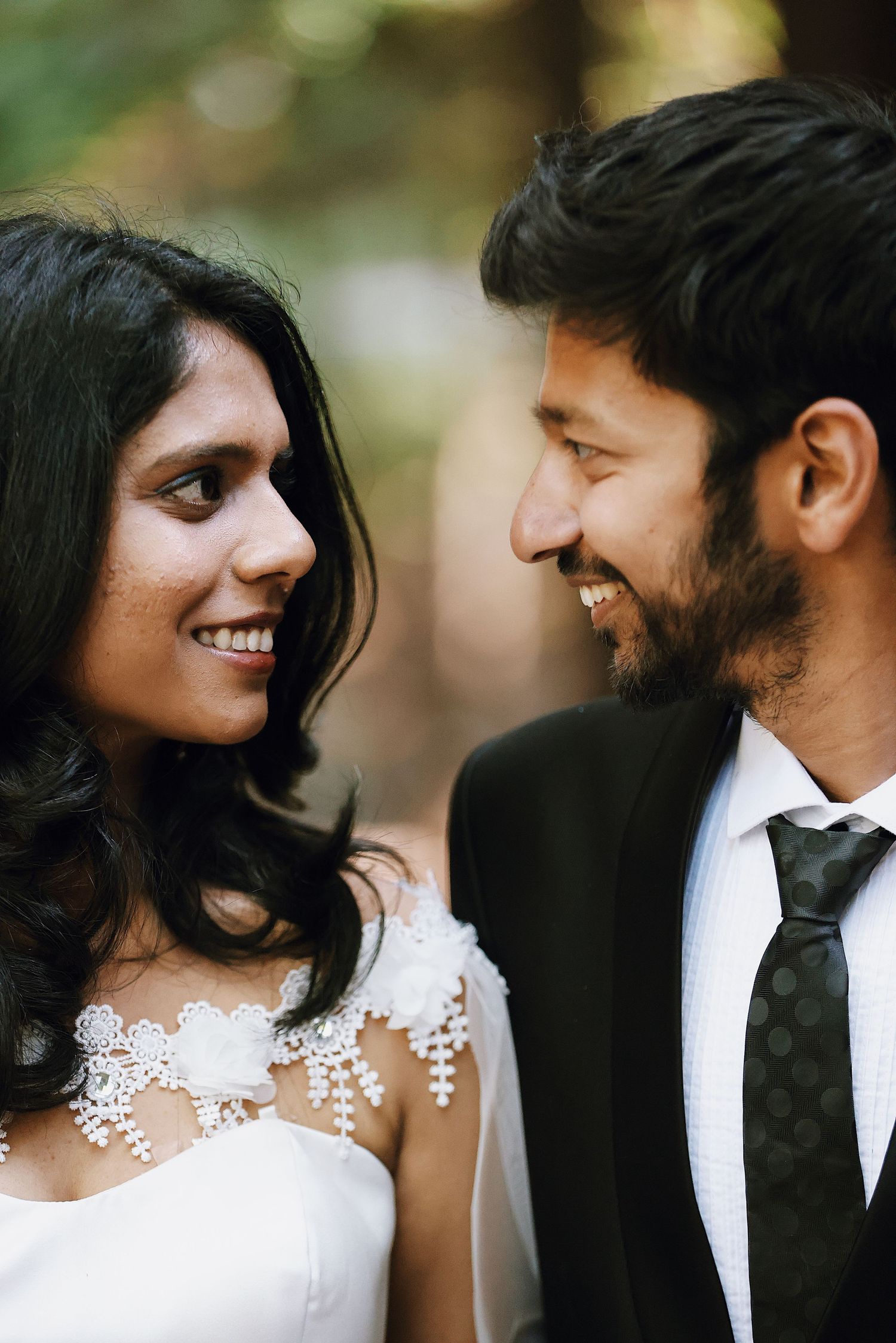 Indian wedding couple smiling at each other, bride in white lace dress and groom in black suit.