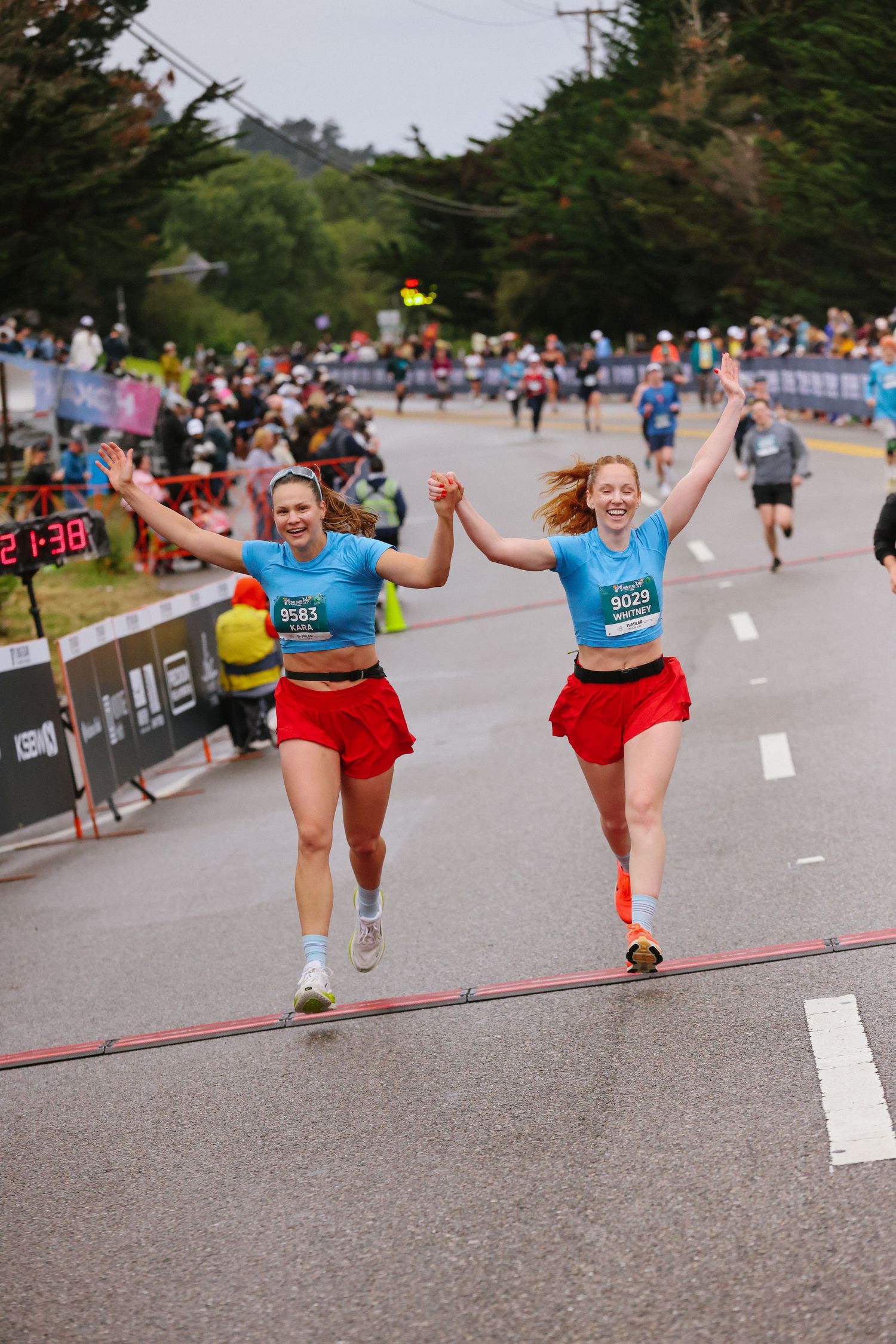 Two women in matching blue tops and red shorts celebrate crossing the finish line of a road race holding hands.