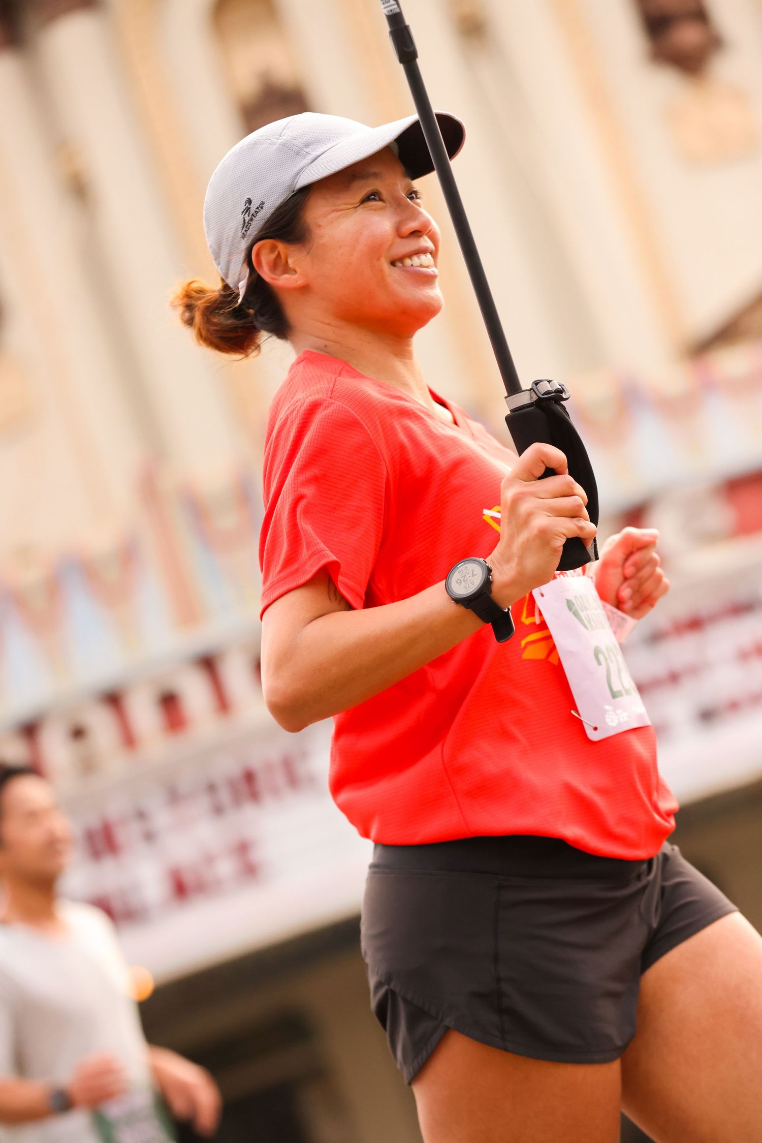A smiling Asian woman in red shirt runs while holding a water bottle at a marathon.