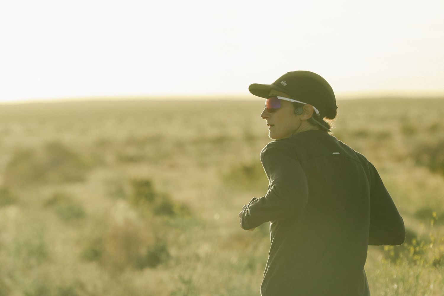 Lone runner in dark jacket and cap jogs through an open sunlit field during golden hour outdoors.