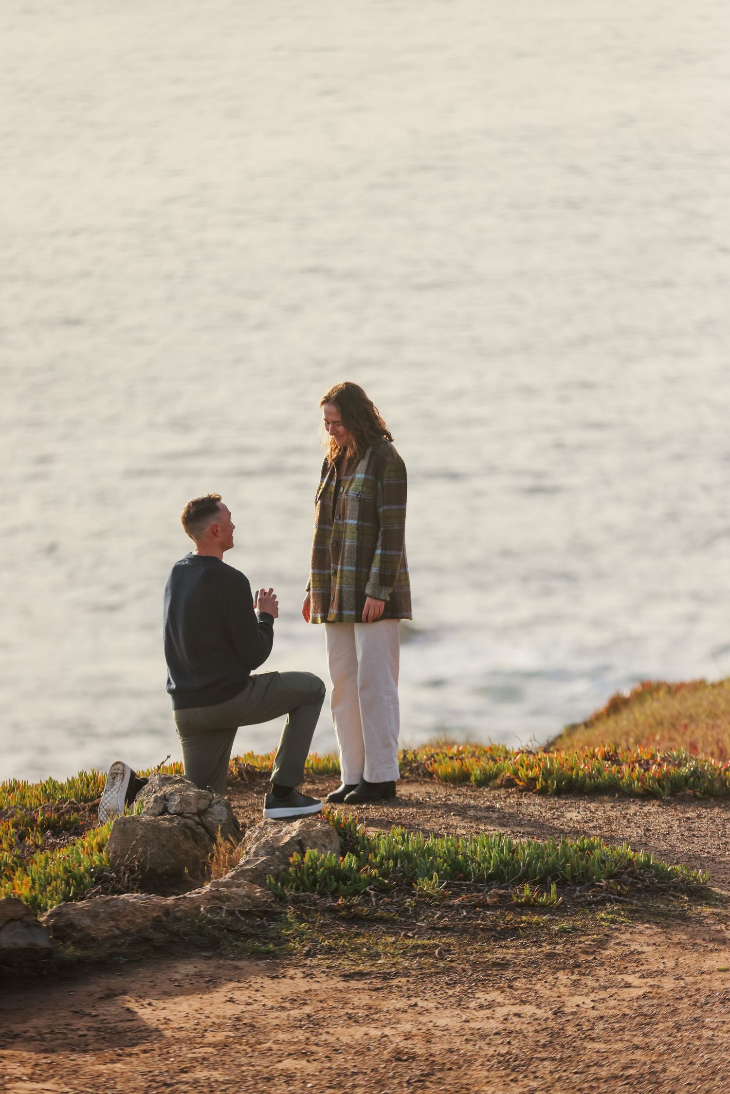 Man proposing on one knee to woman on coastal cliff at sunset with ocean backdrop.