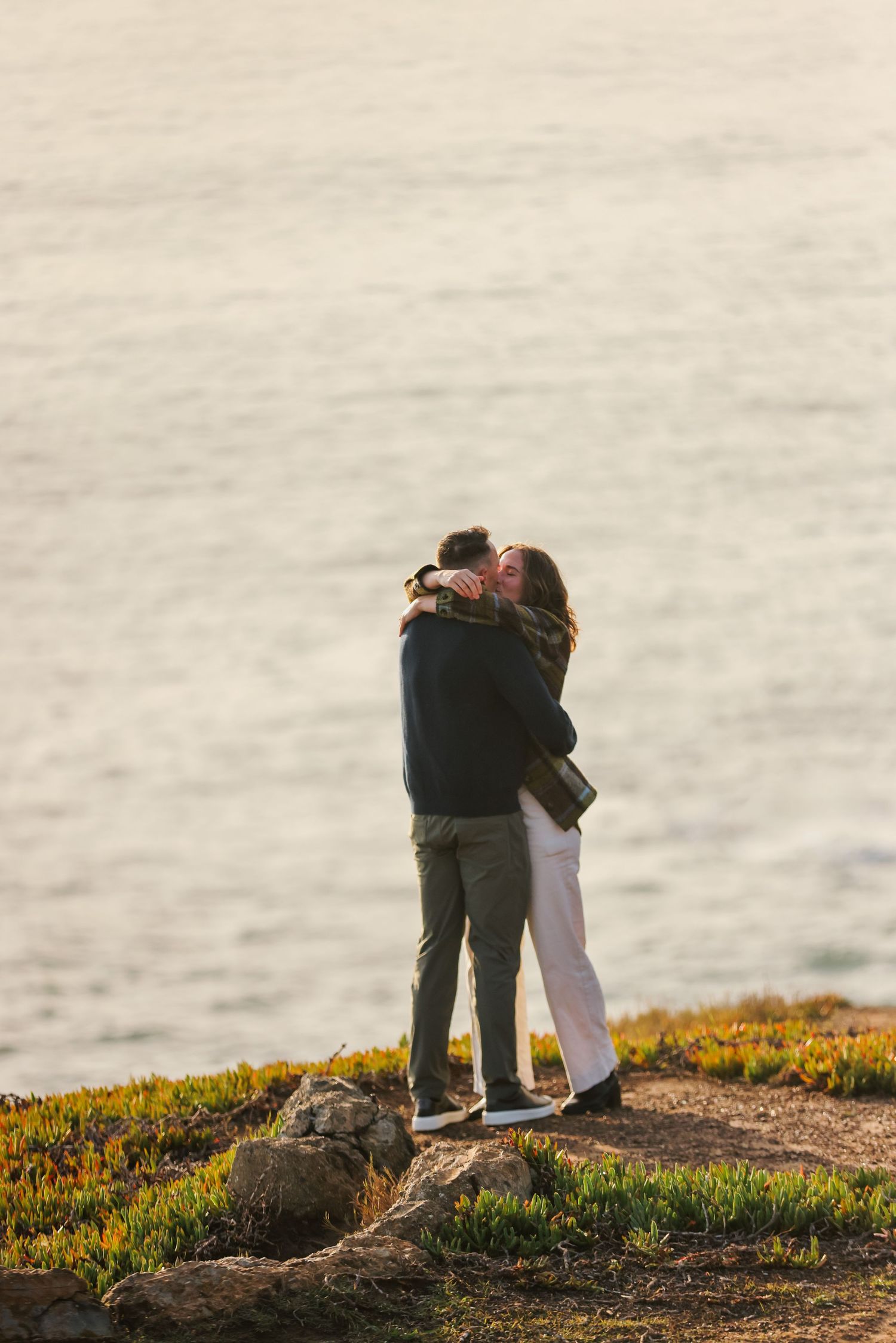 Couple embracing and kissing on coastal cliff at golden hour with sparkling ocean in background.
