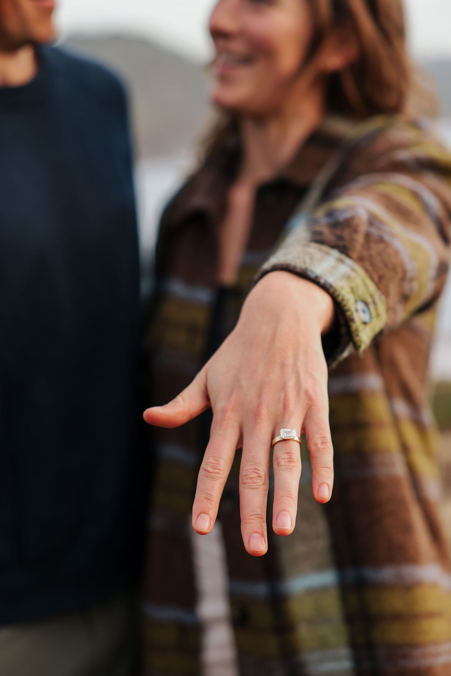 Woman extending hand to show emerald-cut diamond engagement ring outdoors.