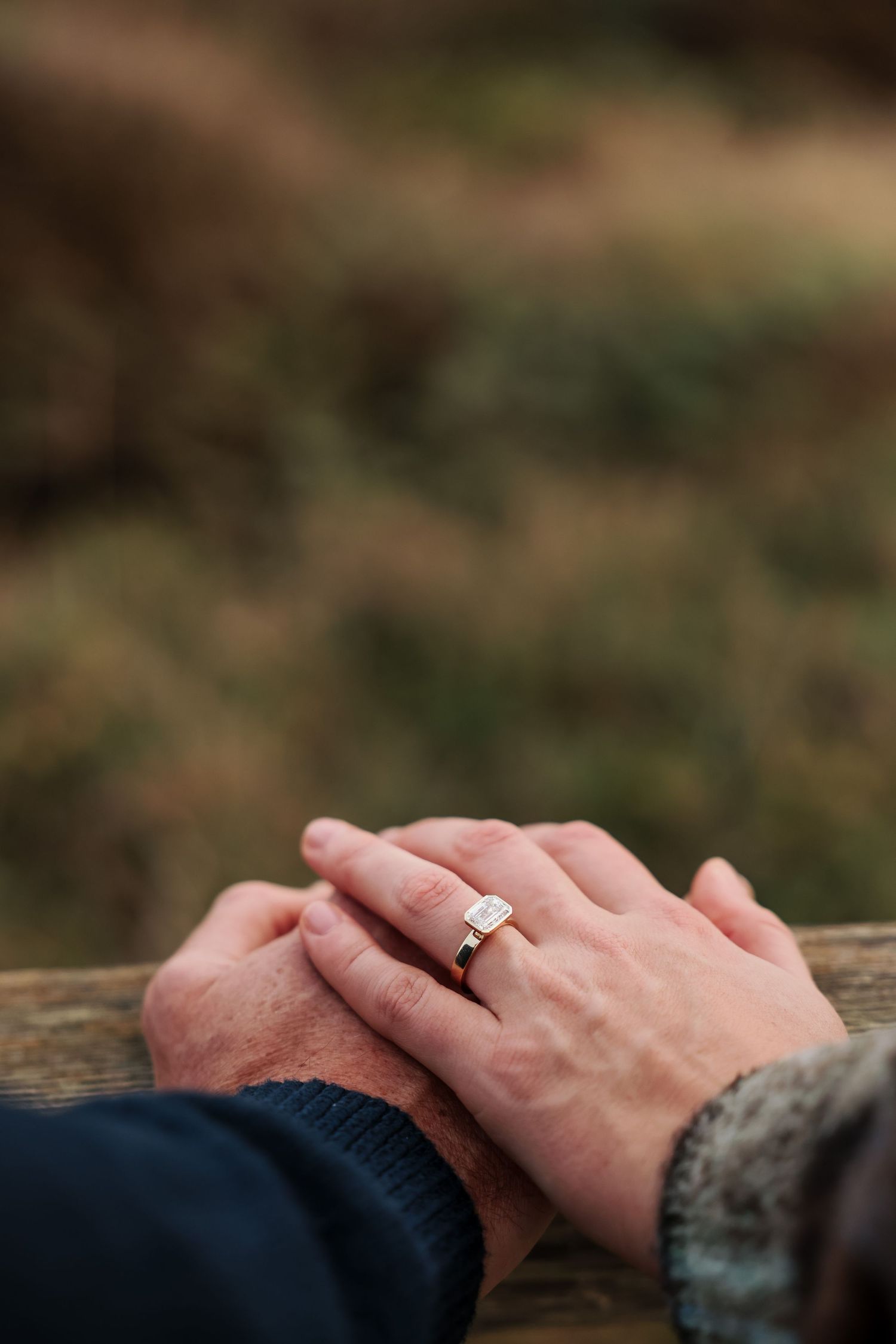 Close-up of engaged couple holding hands showing emerald-cut diamond engagement ring on wooden fence.