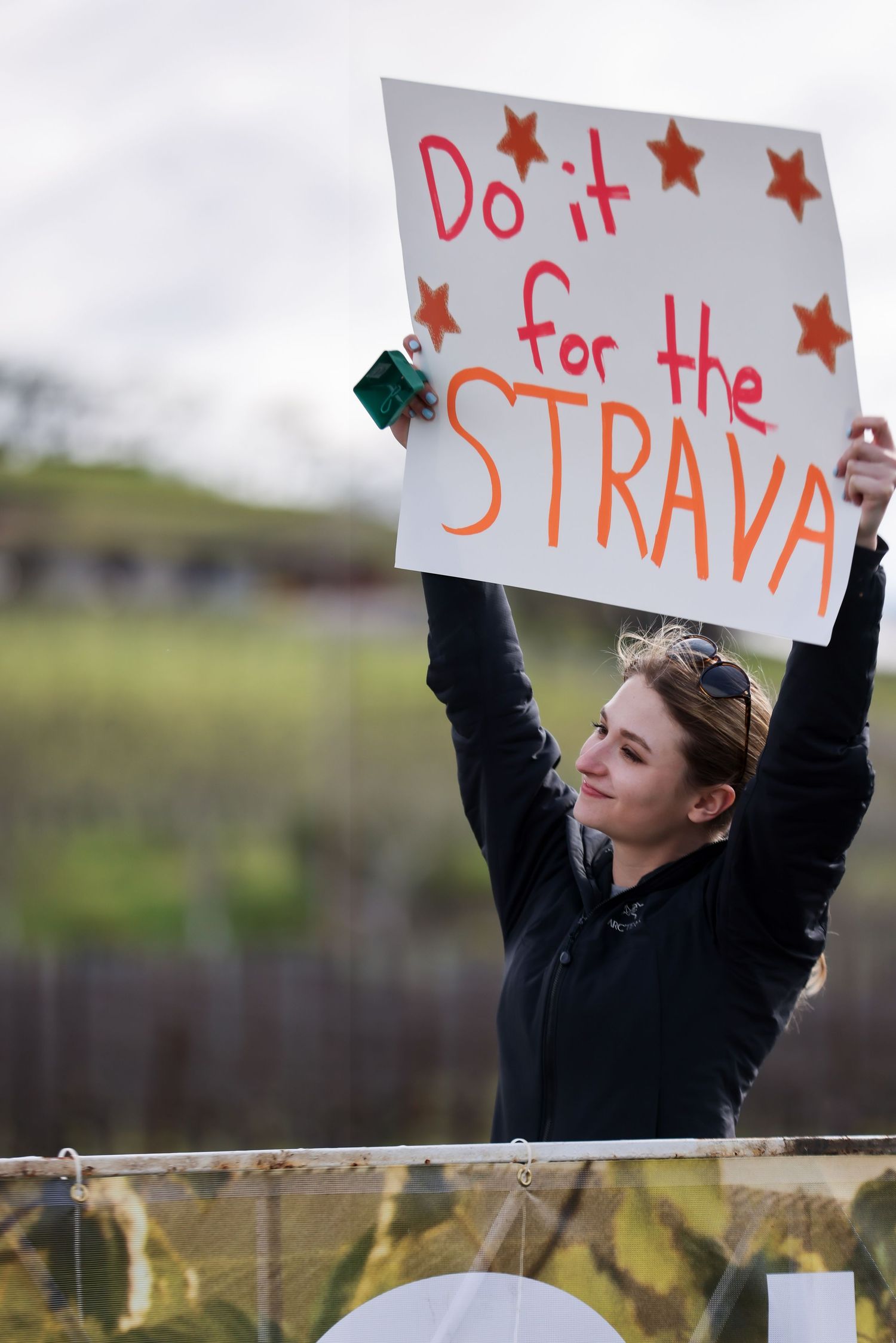 A spectator holds a humorous sign reading Do it for the STRAVA at a running race.