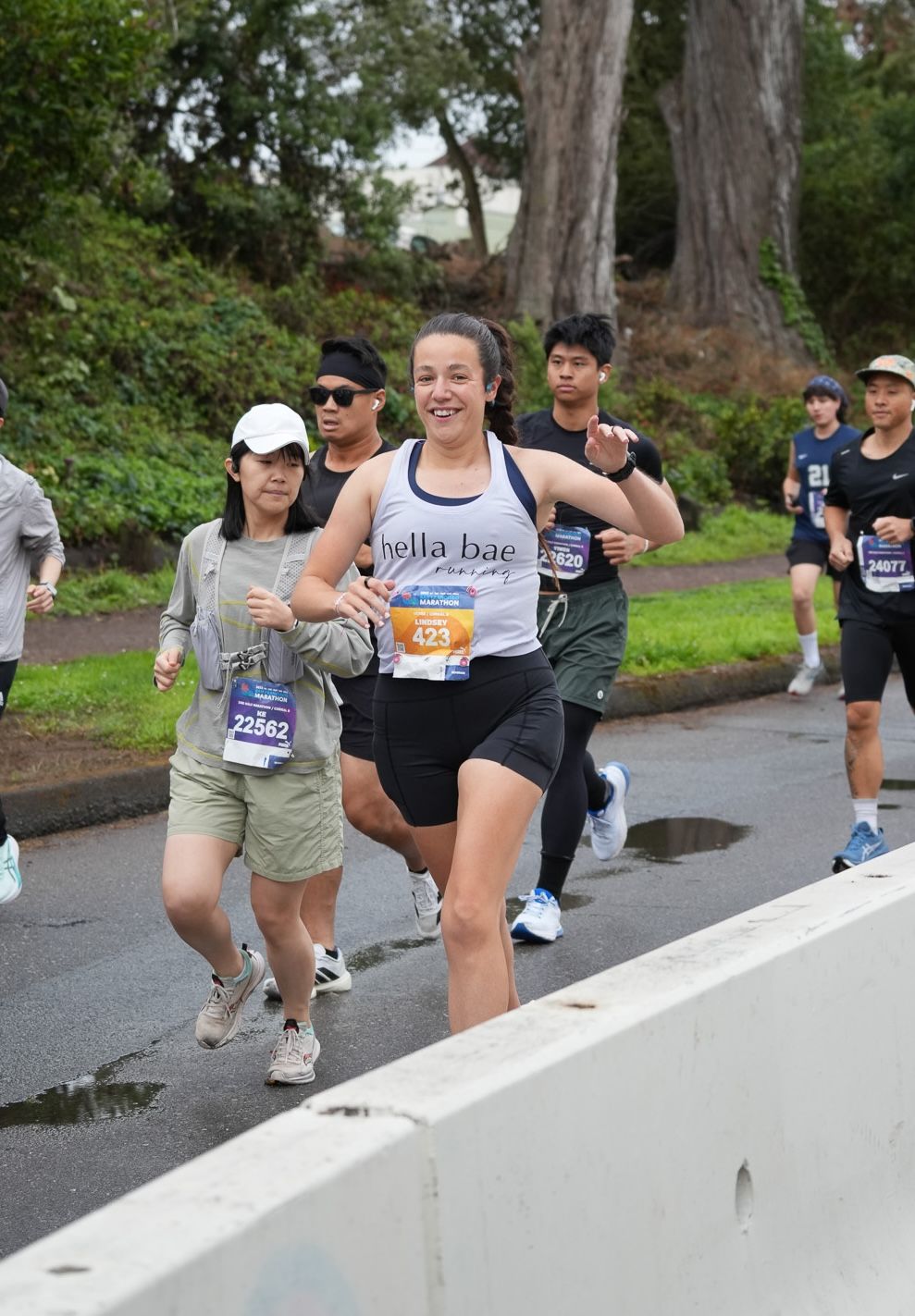 Runners competing in a road race on a wet street, with a woman in a Hella Bae tank top and bib number 425 leading.