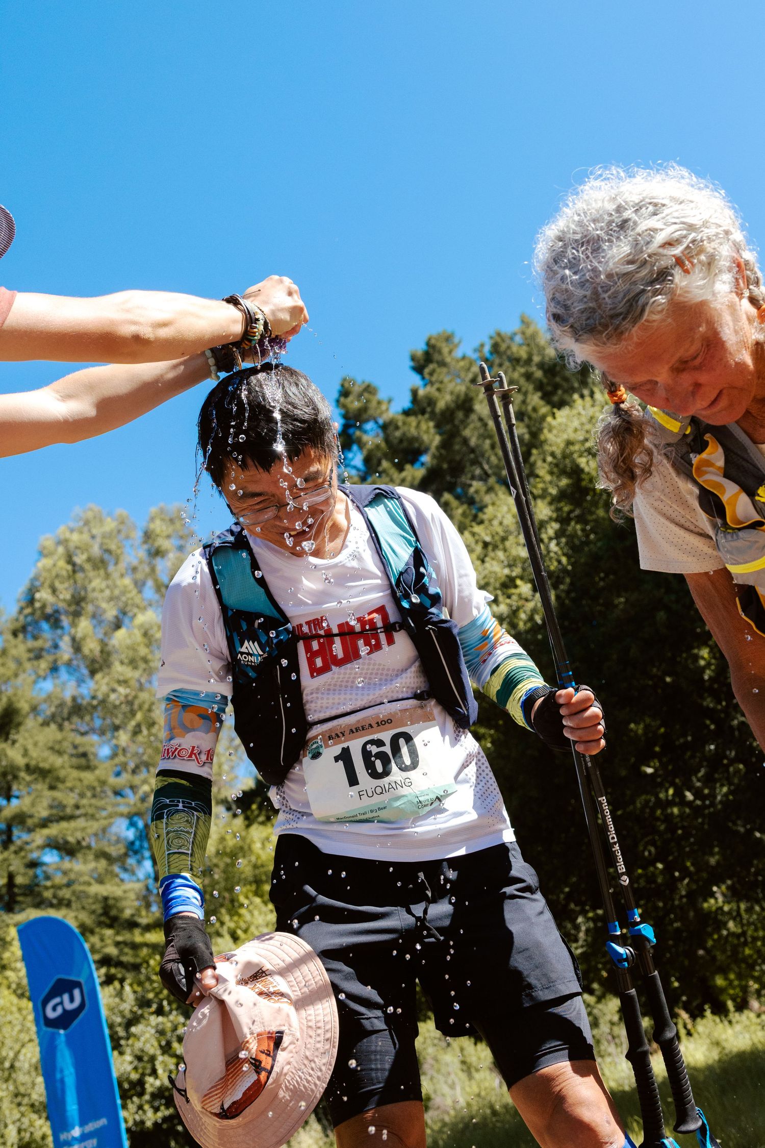 Trail runner #160 gets water poured over head at aid station during sunny ultramarathon race event.