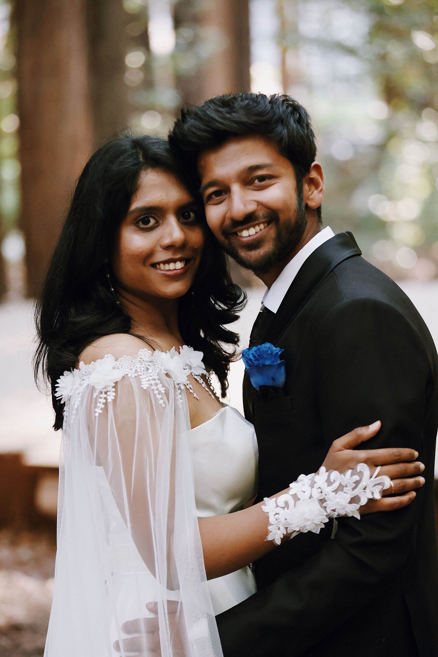 Indian bride and groom embracing outdoors, bride in white lace wedding dress with blue boutonniere groom.