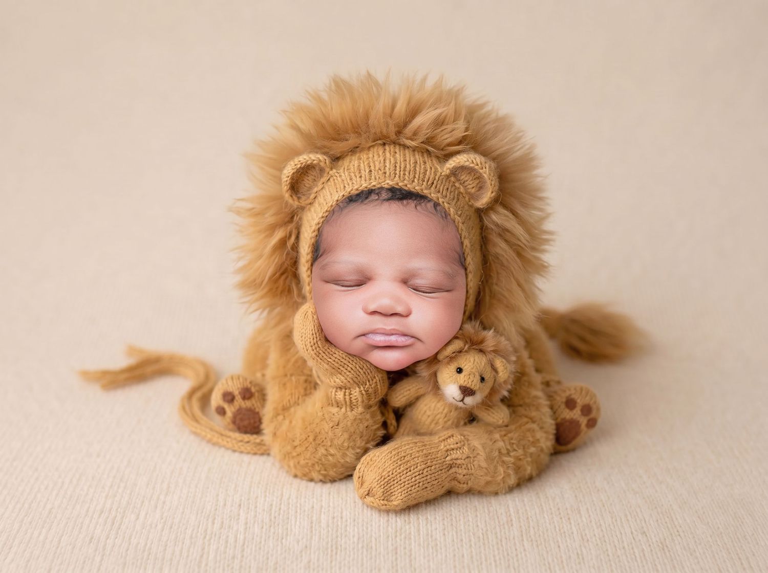 Sleeping newborn dressed in a cozy lion costume cuddles a matching stuffed lion toy on beige backdrop.