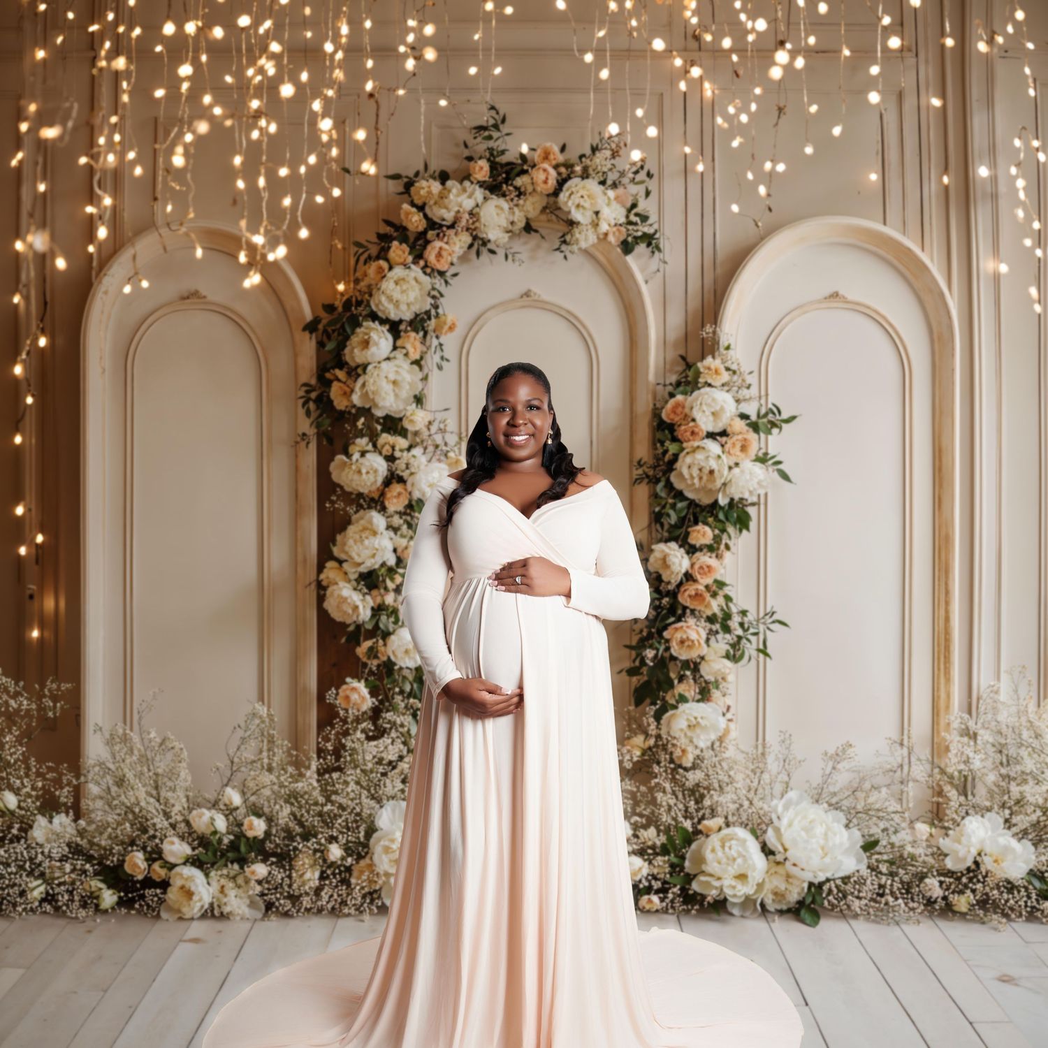 Pregnant woman in elegant white maternity gown poses before a floral arch with warm fairy lights.