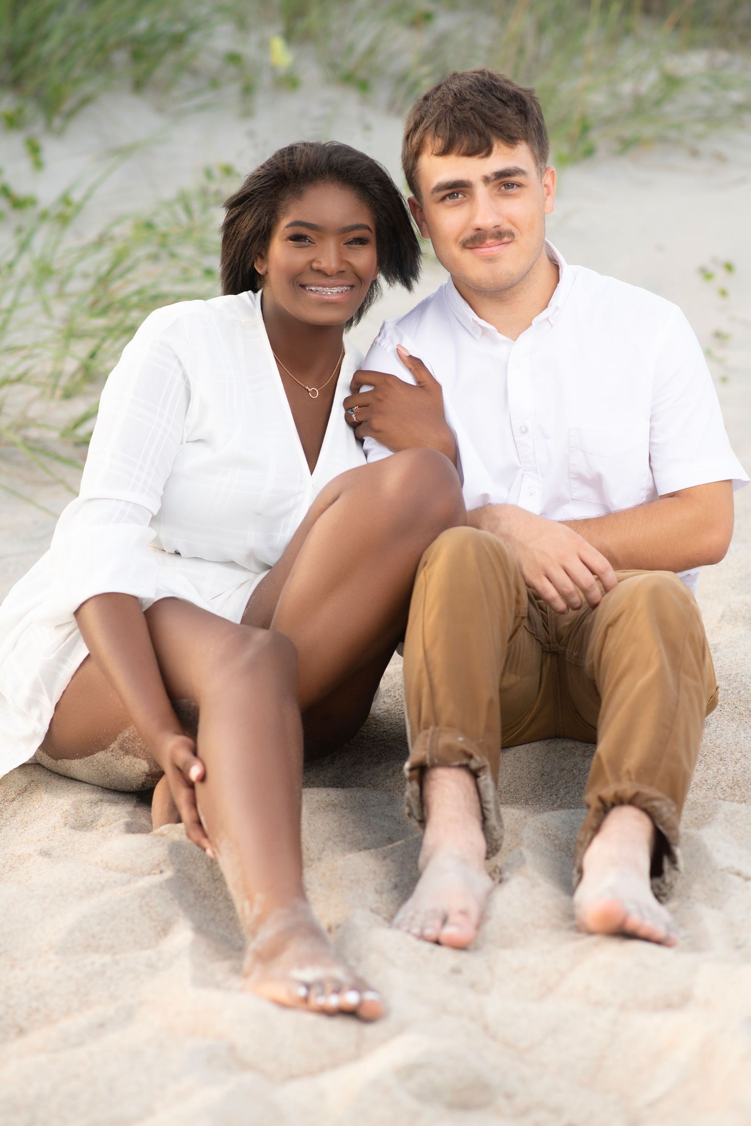Interracial couple in white outfits sit together barefoot on sandy beach dunes smiling warmly.