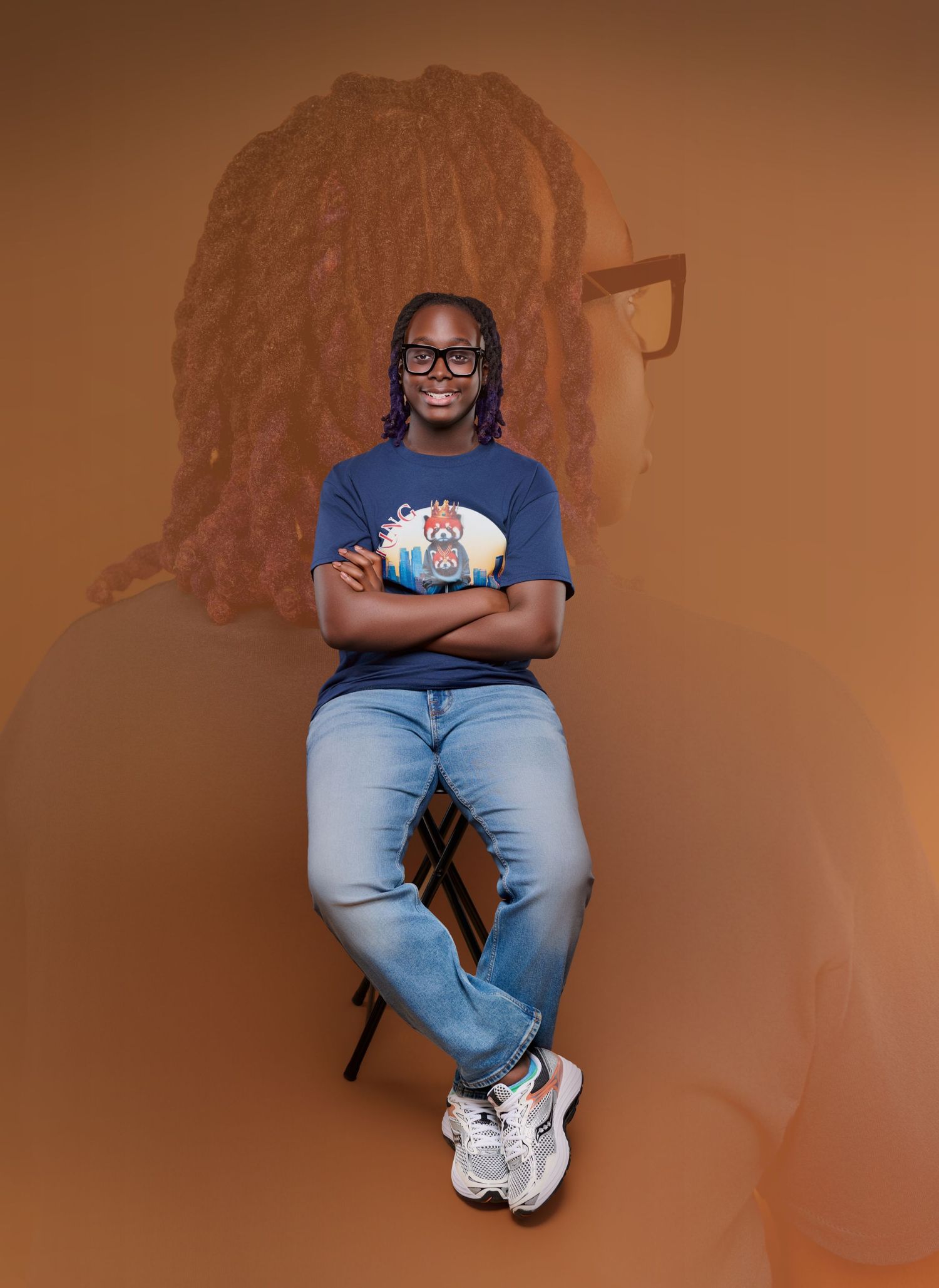 Young man with locs in blue graphic tee and jeans sits confidently against an orange toned background.