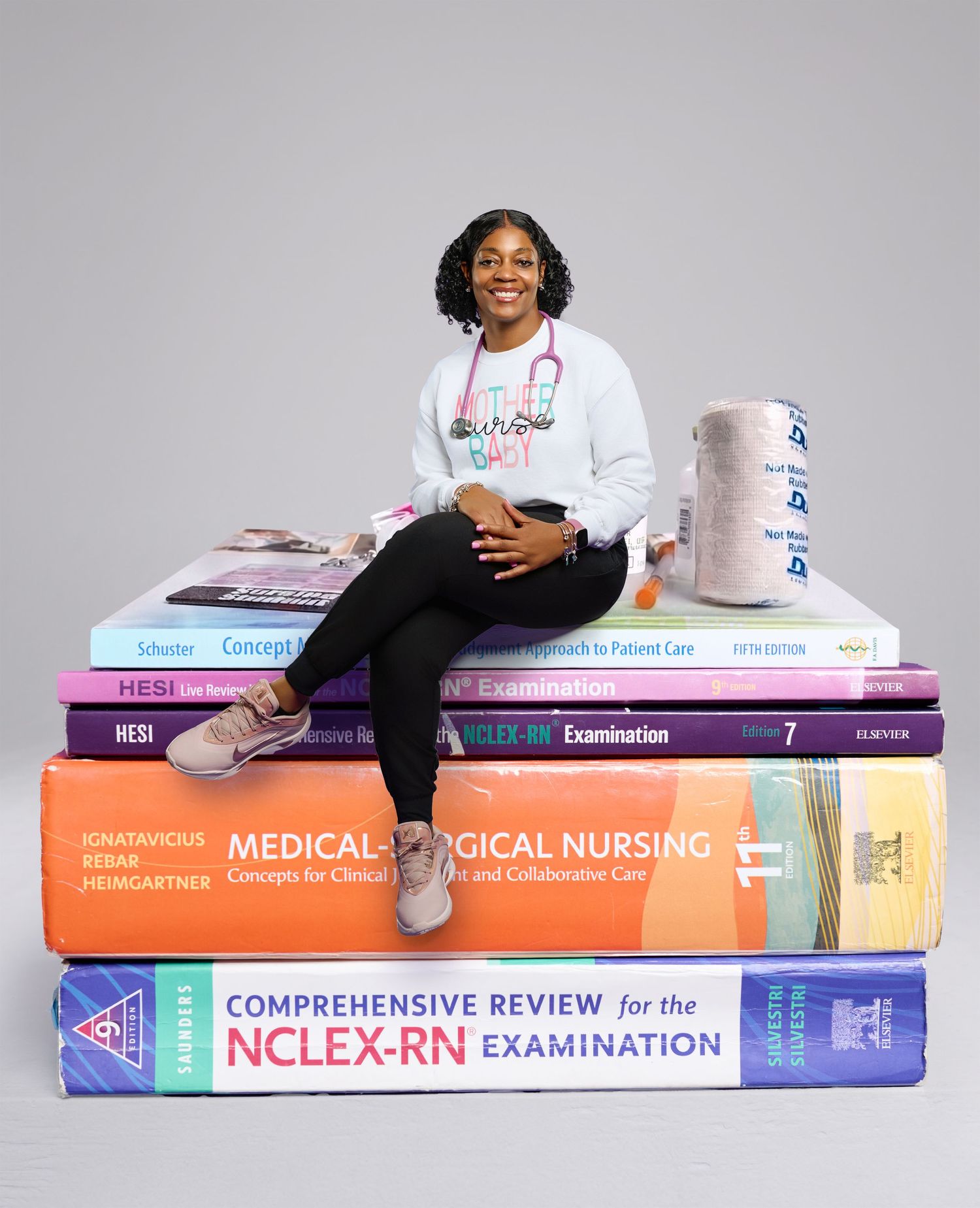 Nursing student sitting atop stack of medical textbooks including NCLEX-RN review books, wearing nurse sweatshirt.