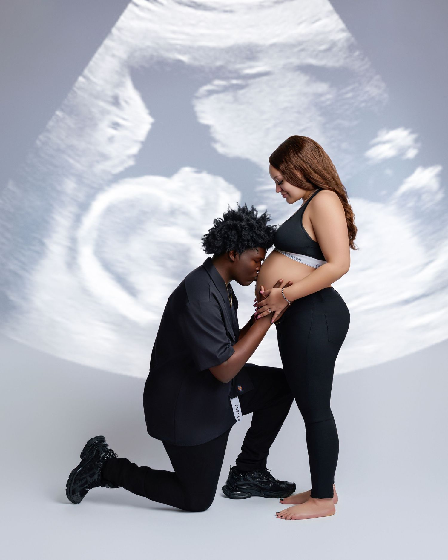 Couple posing with ultrasound image backdrop, man kneeling kissing pregnant womans belly in black outfits.