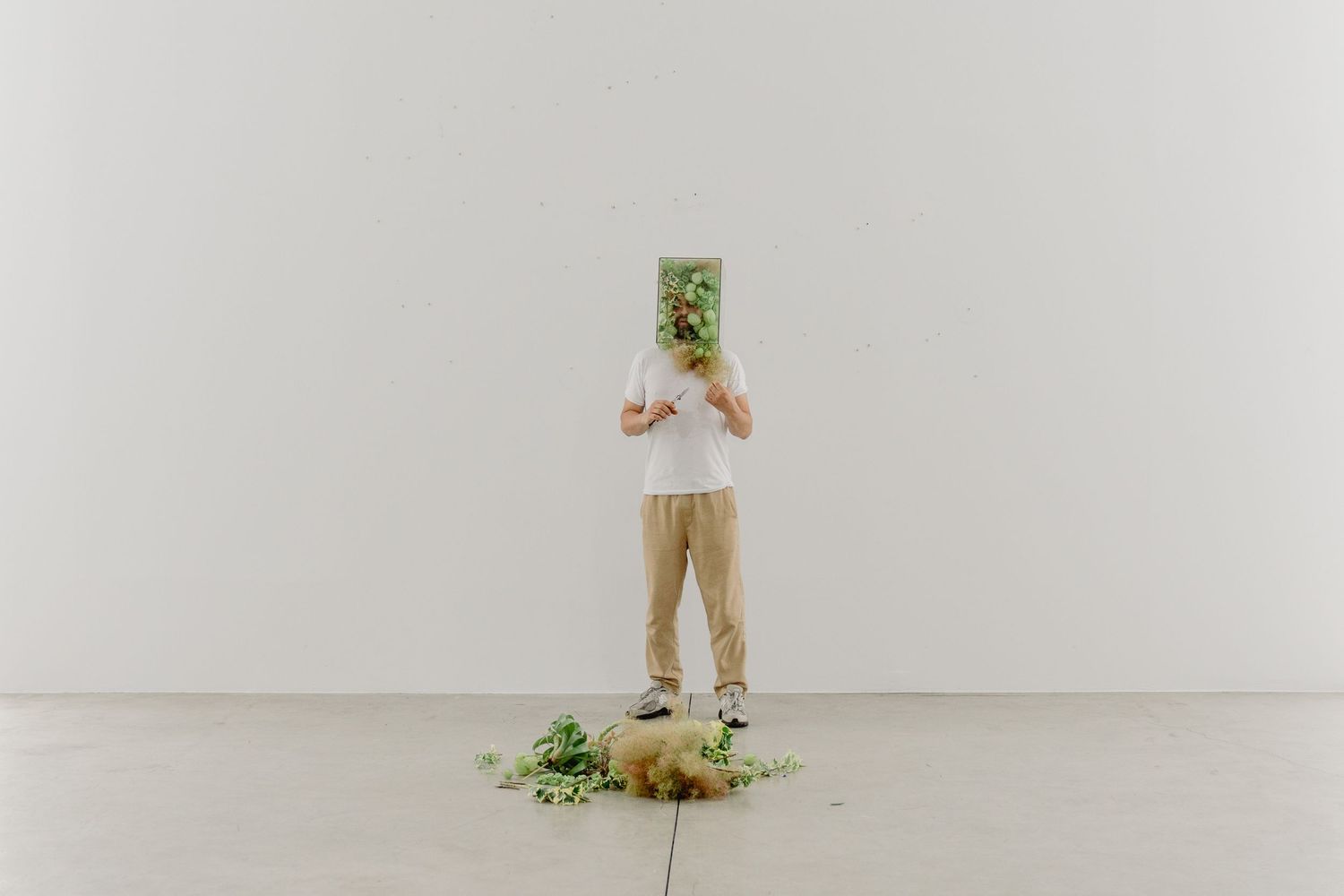 The Florist Artist Yunus Karma holding green plant box over face, standing on white floor with scattered leaves in minimalist gallery space.