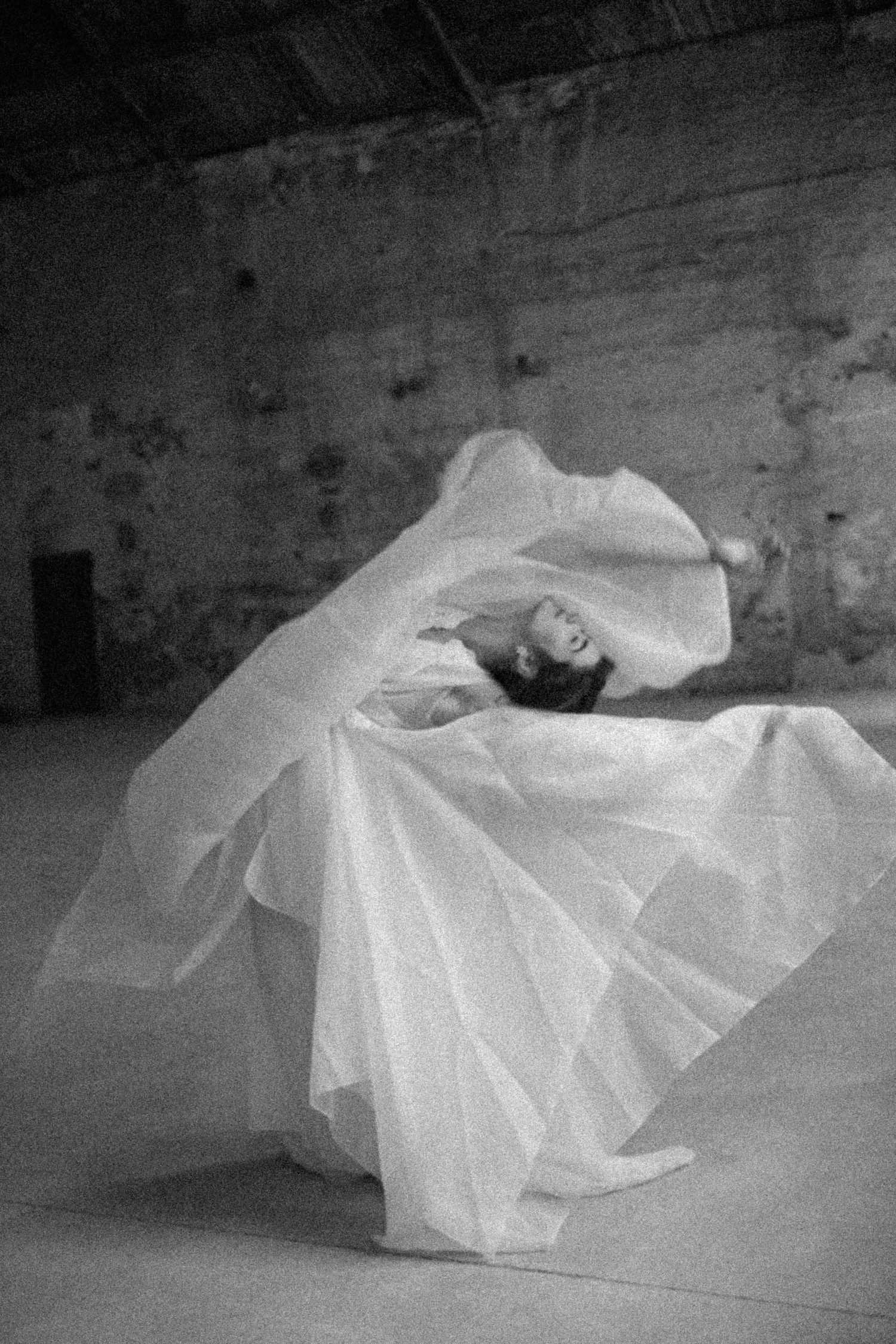 Black and white photo of a  bride in a flowing white gown twirling gracefully in a rustic stone room.