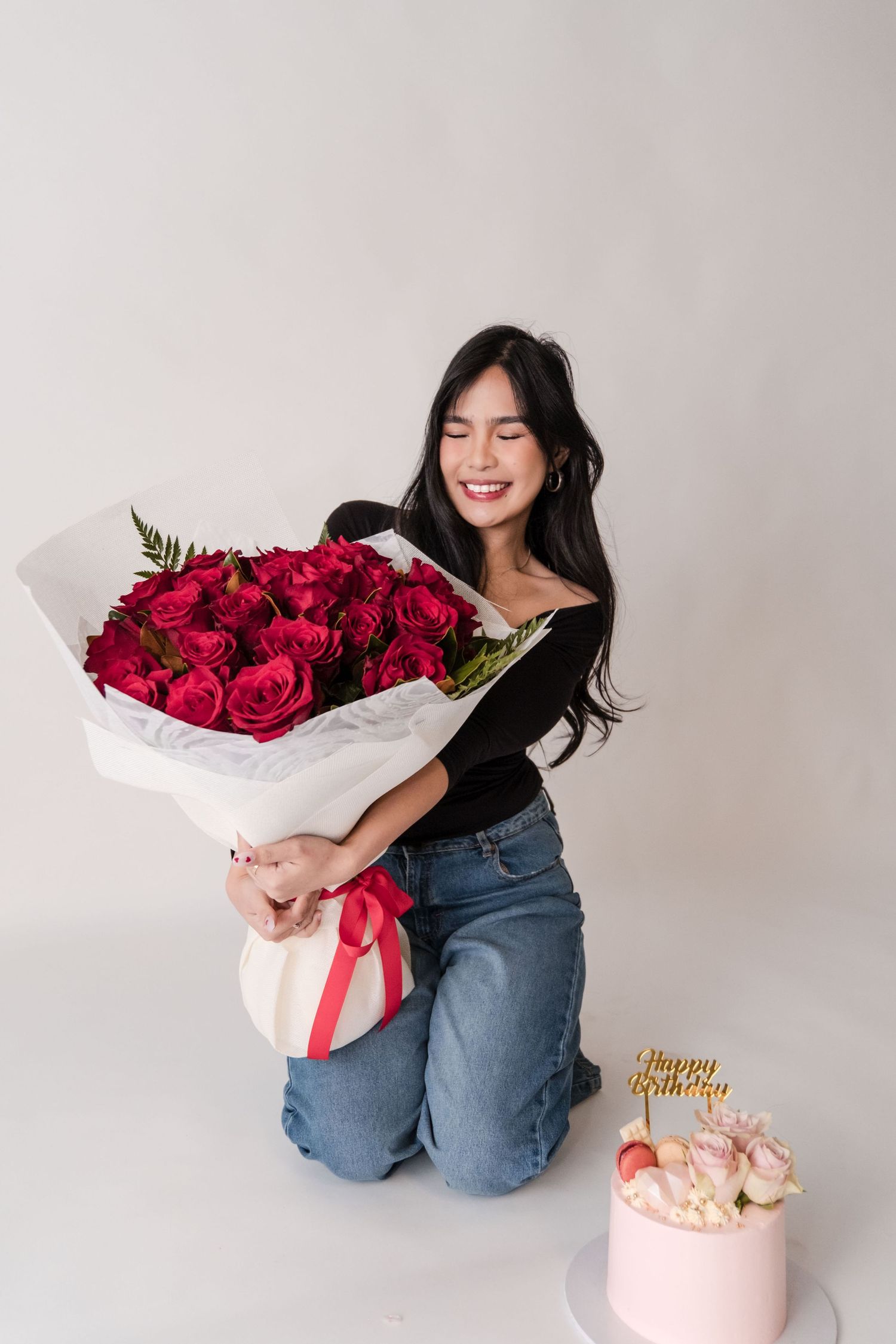 Smiling woman kneeling with large red rose bouquet beside a Happy Birthday cake on white background.