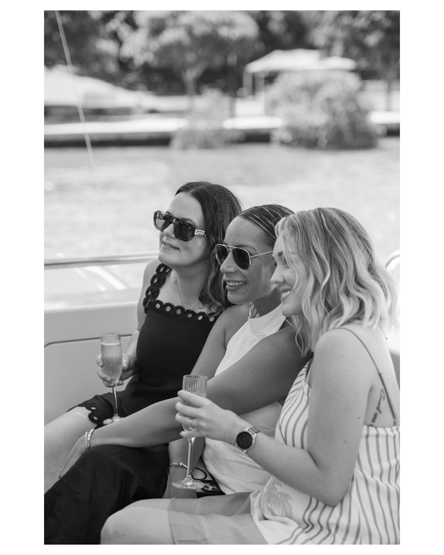 Three women in sunglasses enjoying champagne on a boat, smiling together on a sunny day.