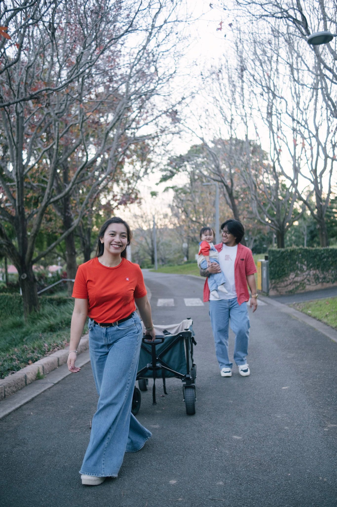 Family strolling in a tree-lined park, woman in red top pulling wagon, another carrying toddler in blue jeans.