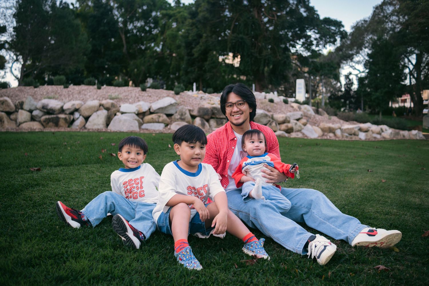 A man sits on green grass with three young children, smiling outdoors near a stone wall and trees.