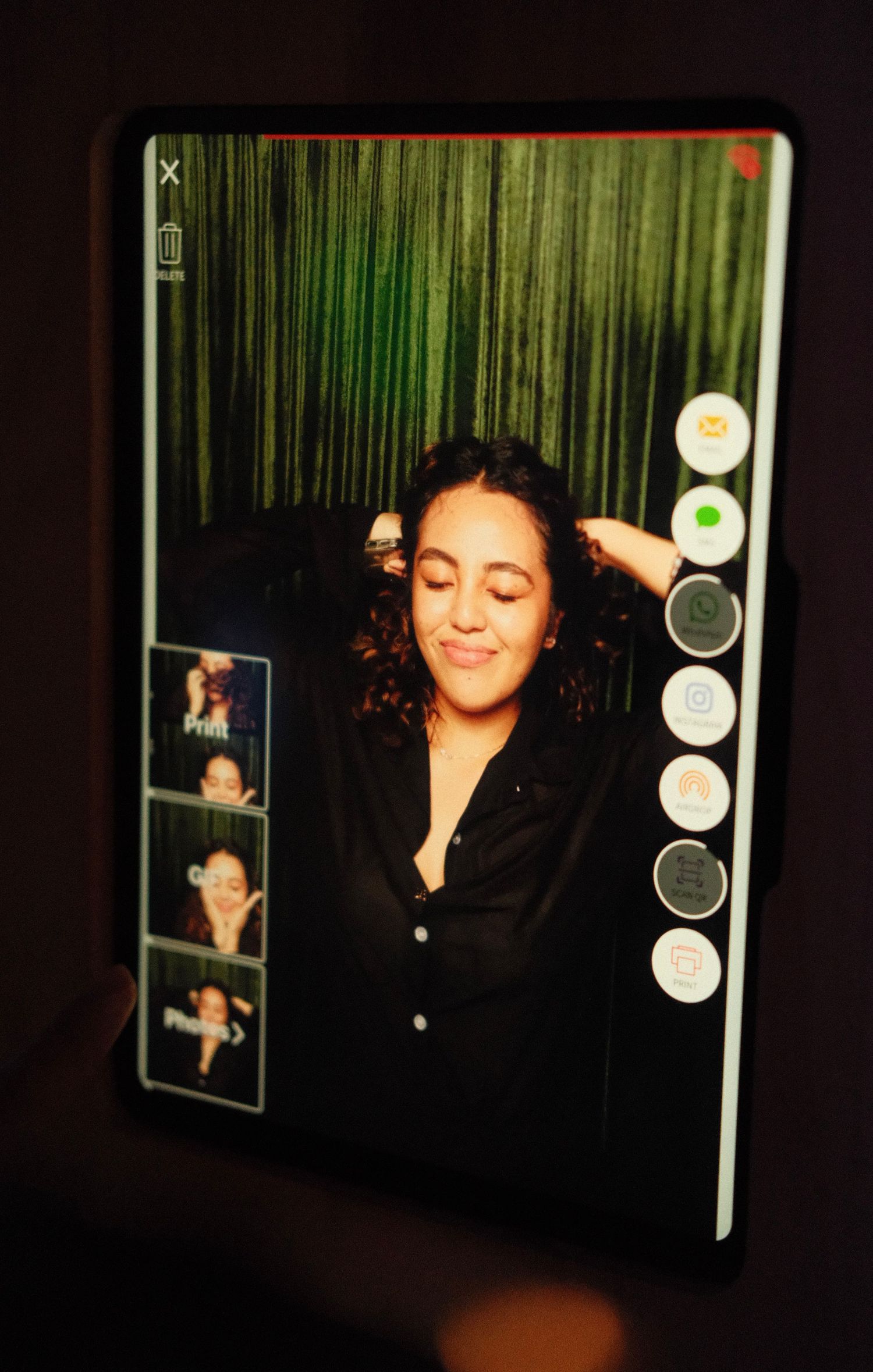Woman posing in a photo booth with curly hair, black shirt, and green curtain backdrop displayed on a tablet screen.