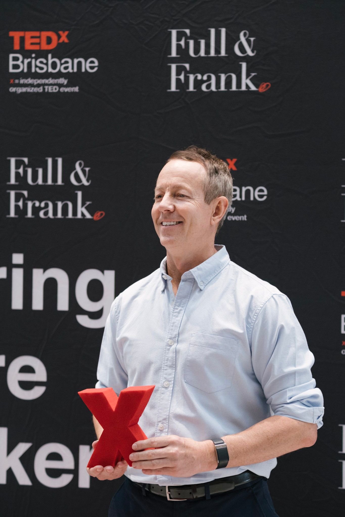 Man holding red TEDx logo at TEDxBrisbane event with Full & Frank sponsor backdrop.