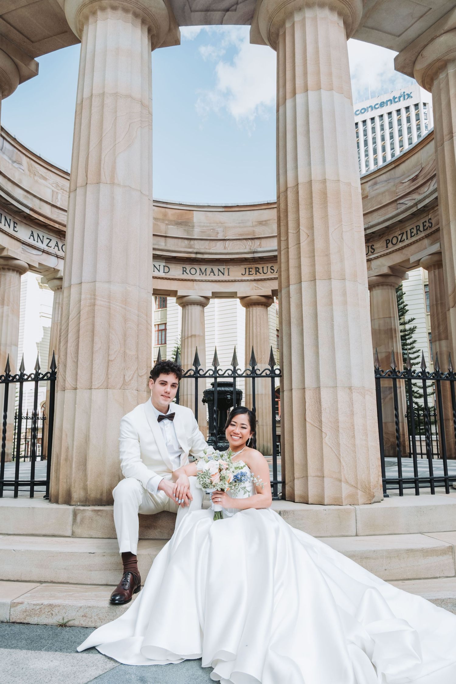 Bride and groom pose among grand stone columns at a classical monument, bride holding a white bouquet in full wedding attire.