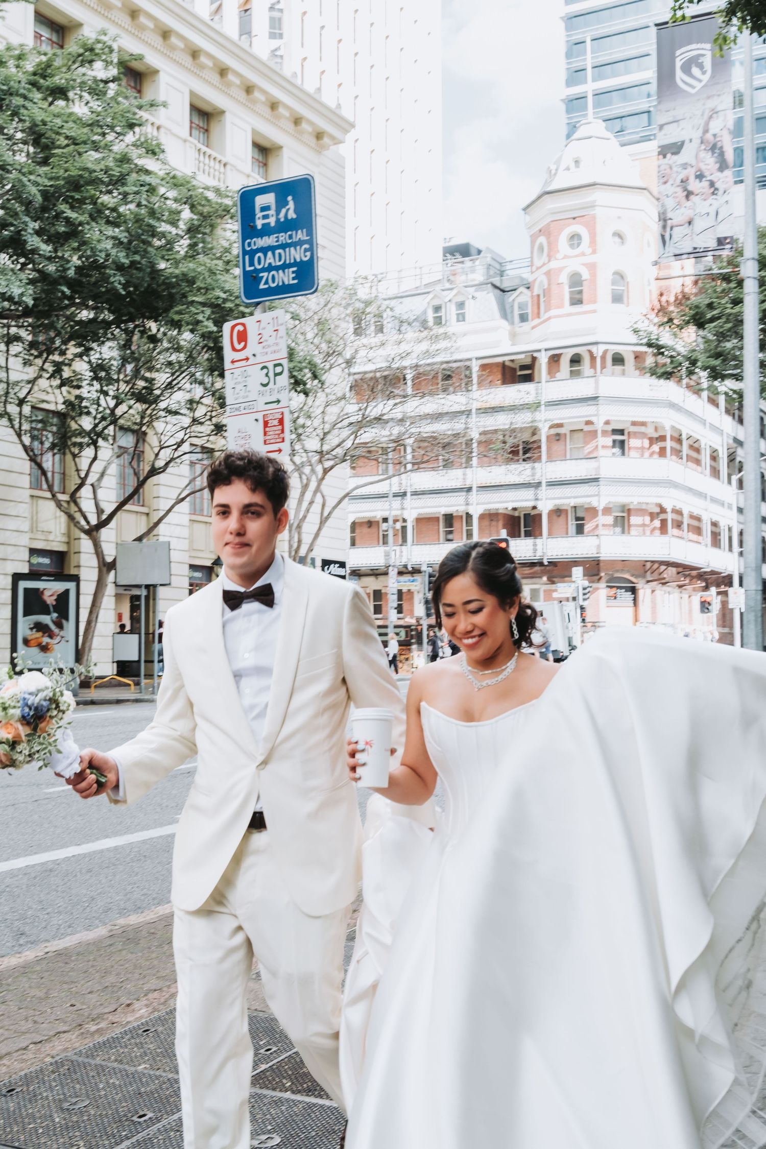 Bride and groom in white wedding attire walking through city streets, bride holding coffee cup near commercial loading zone sign.