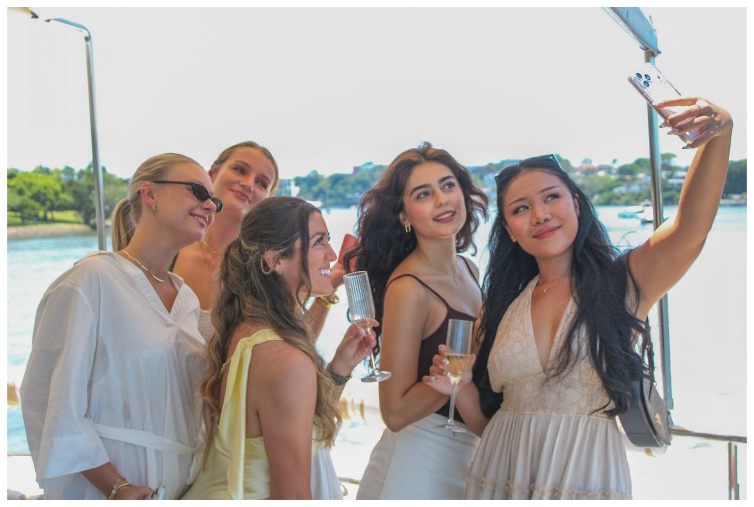 Five women taking a selfie on a boat, holding champagne glasses and smiling outdoors.