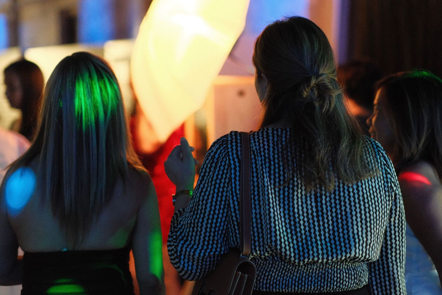 Two women viewed from behind at a colorful evening event with glowing lights in blue and green.