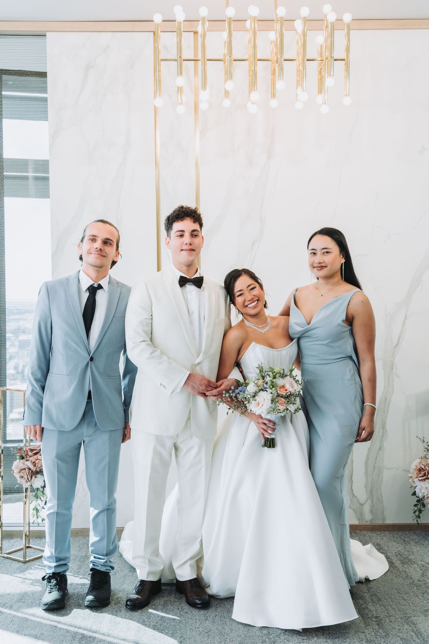 Wedding party of four posing indoors, bride in white gown with bouquet, groom in cream tuxedo, guests in blue formal attire.