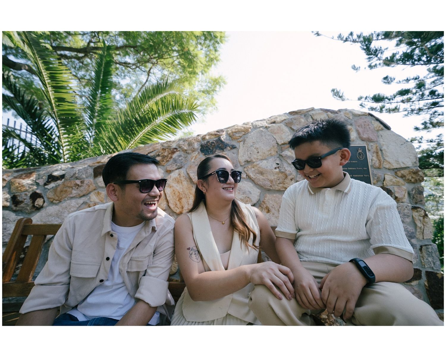 Three friends in sunglasses and beige outfits smile and laugh together outdoors near a stone wall and palm trees.