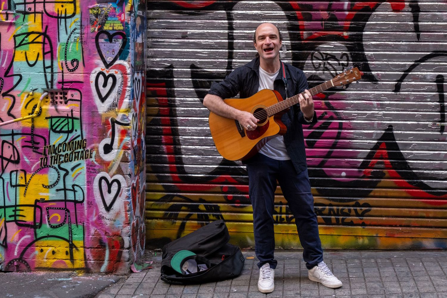 A musician playing an acoustic guitar on a street with graffiti-covered walls.