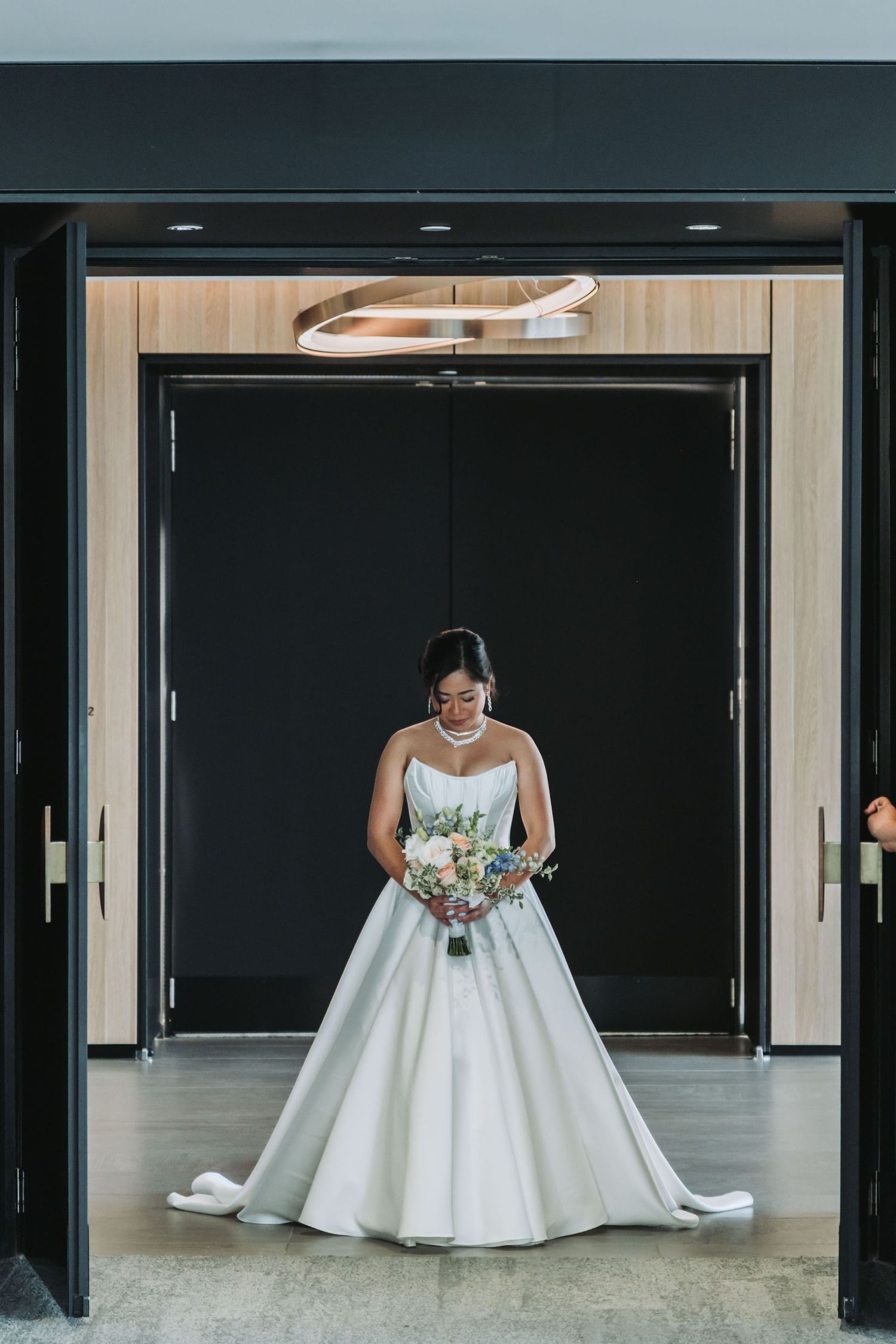 Bride in strapless white ball gown holding bouquet stands in elegant doorway with modern circular chandelier.