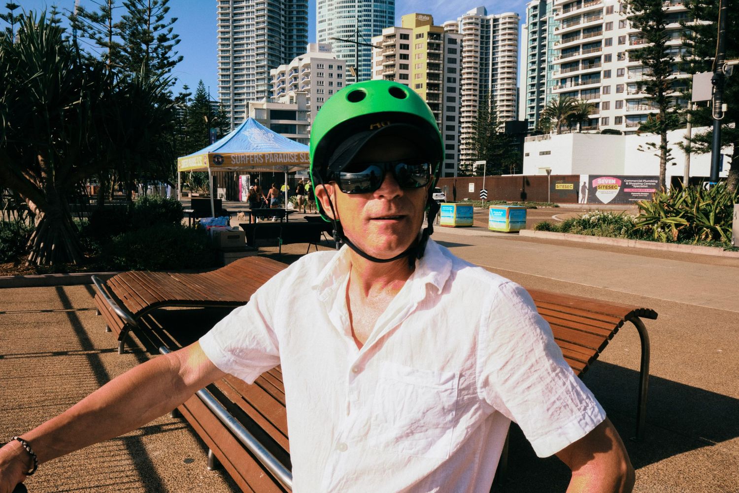 A man wearing a white shirt and a bright green bicycle helmet, sitting on a bench.