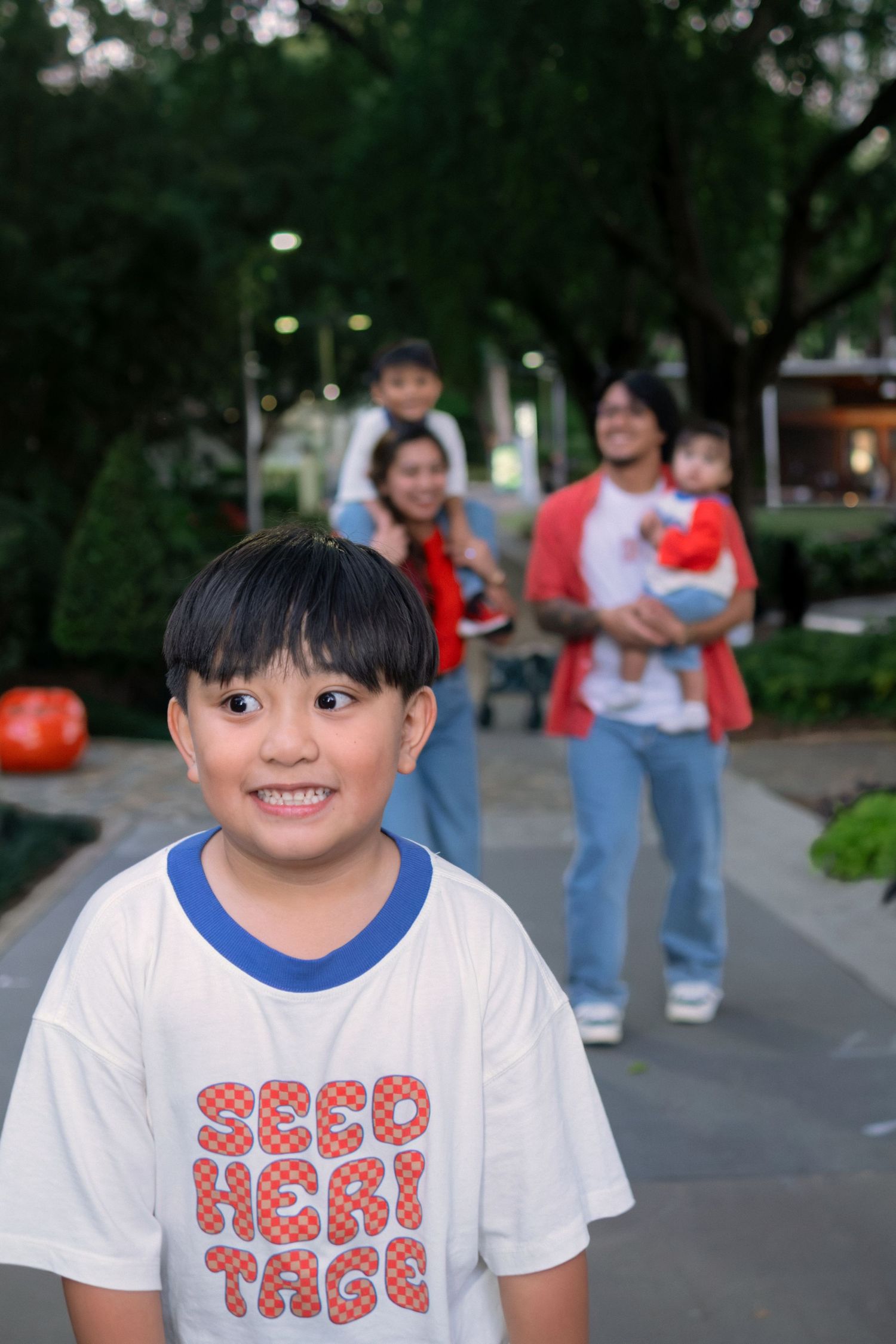 Smiling Asian boy in white Red Heritage t-shirt poses in park with family standing behind him.