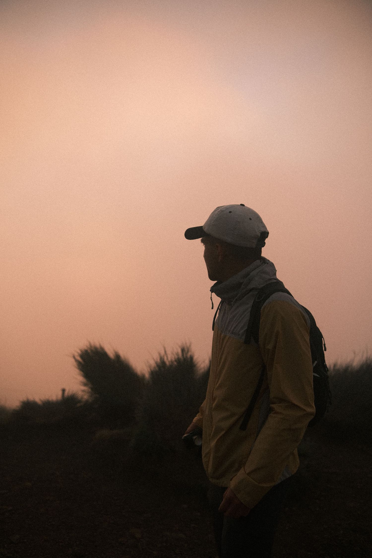 Hiker in yellow jacket and white cap stands against a moody orange dusk sky in a misty landscape.