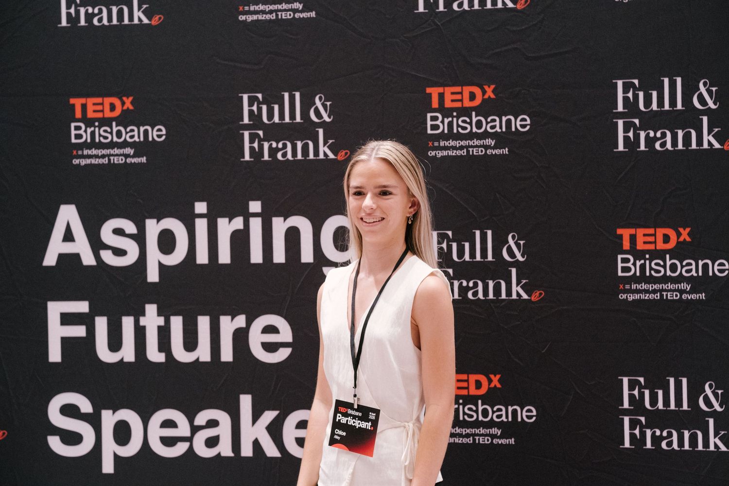 Young woman smiling at TEDx Brisbane Aspiring Future Speakers event, standing in front of Full & Frank branded backdrop.